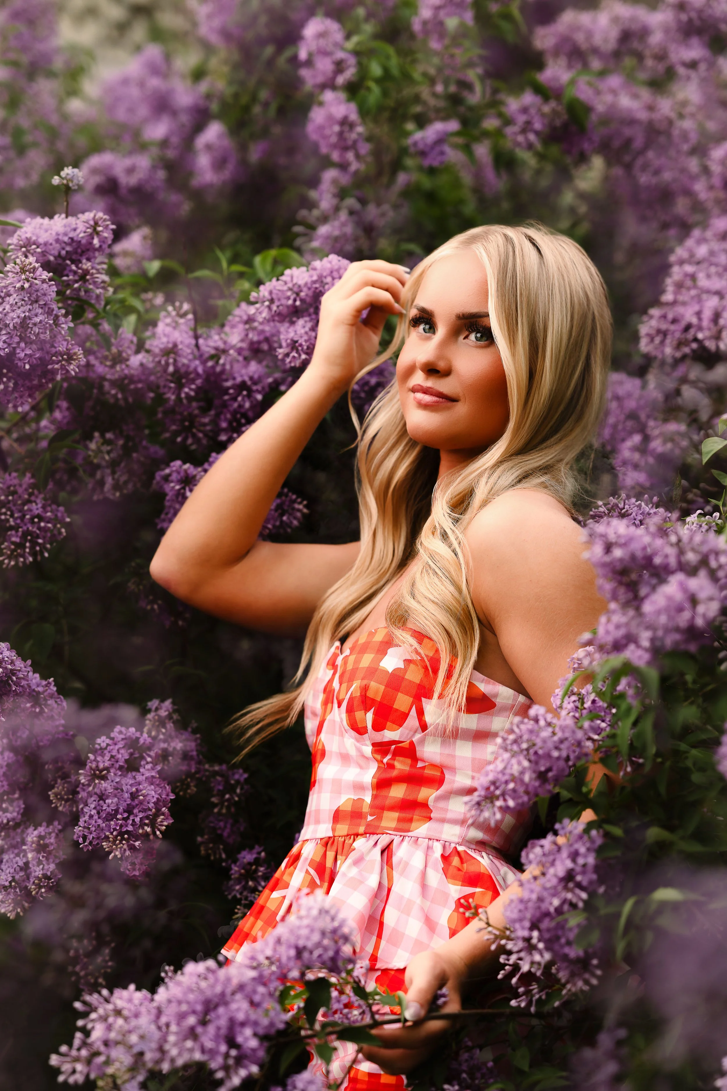A young woman with long blonde hair, dressed in a pink and orange checkered dress, standing among purple lilac flowers. She is looking at the camera with a gentle smile, with her hand touching her hair.