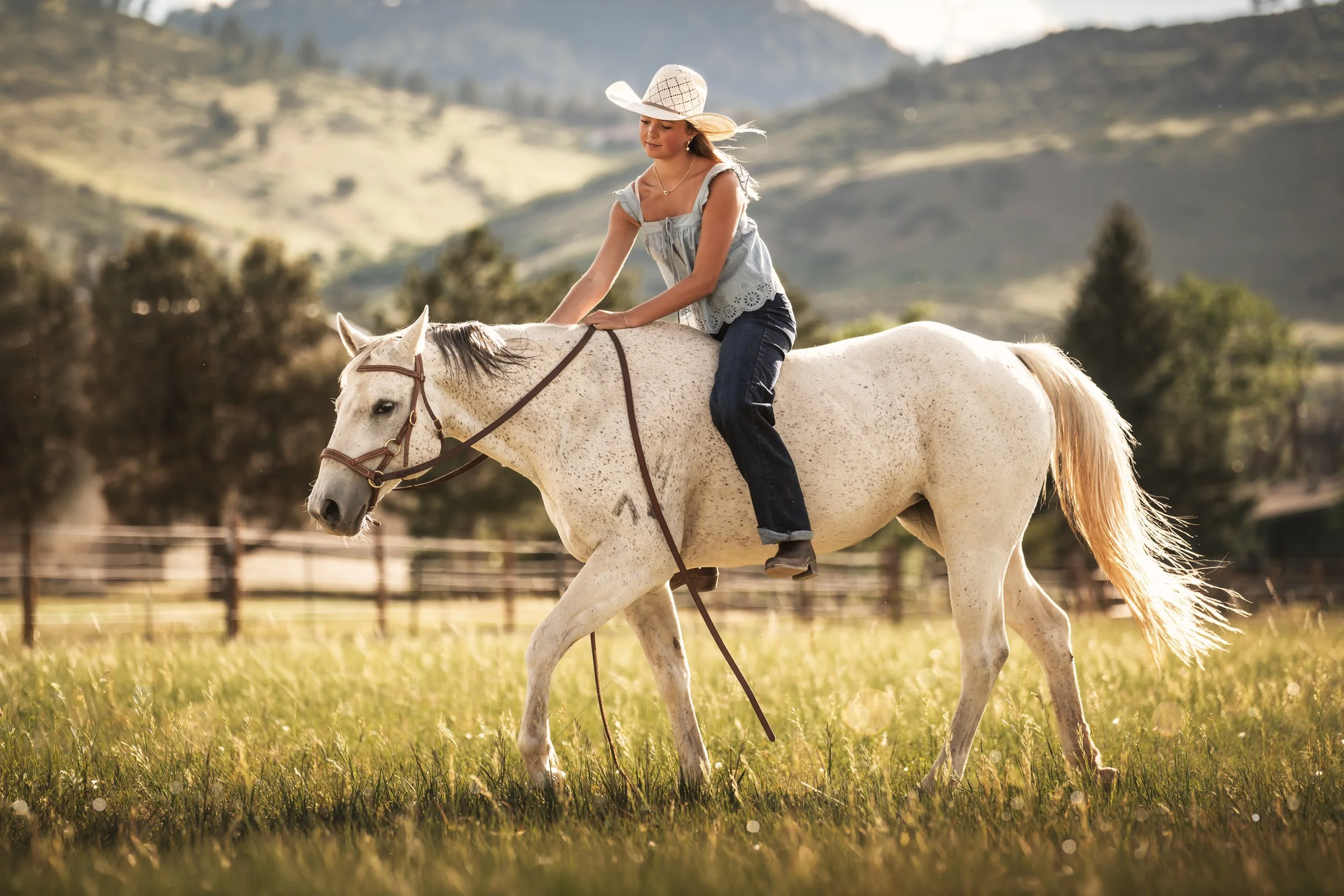 A blonde girl in jeans and a blue tank top with a straw hat rides a white horse in front of the foothills in Boulder Colorado.