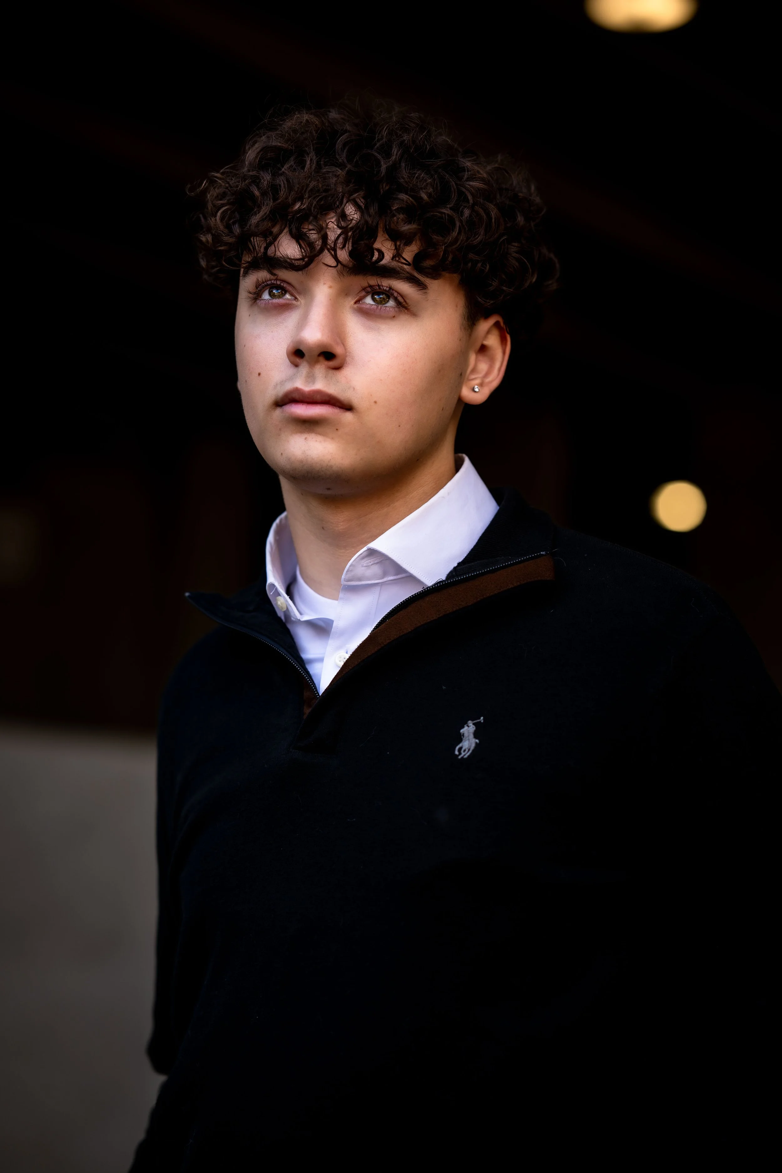A young man with curly hair looking upwards, wearing a white collared shirt and a dark sweater with a small logo, standing in a dimly lit environment.
