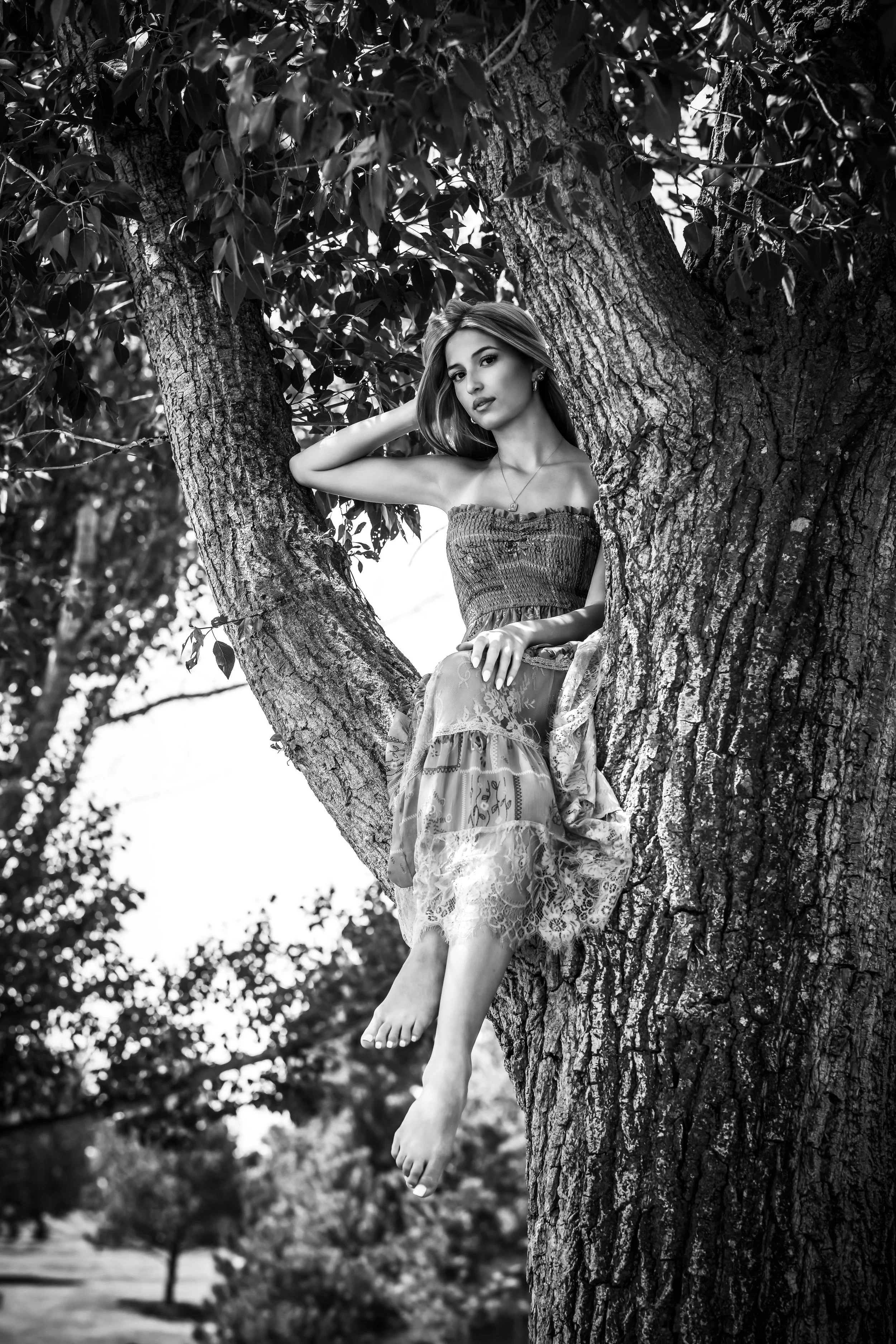 A woman sitting on a tree branch, with her bare feet hanging down, in an outdoor setting with trees in the background.