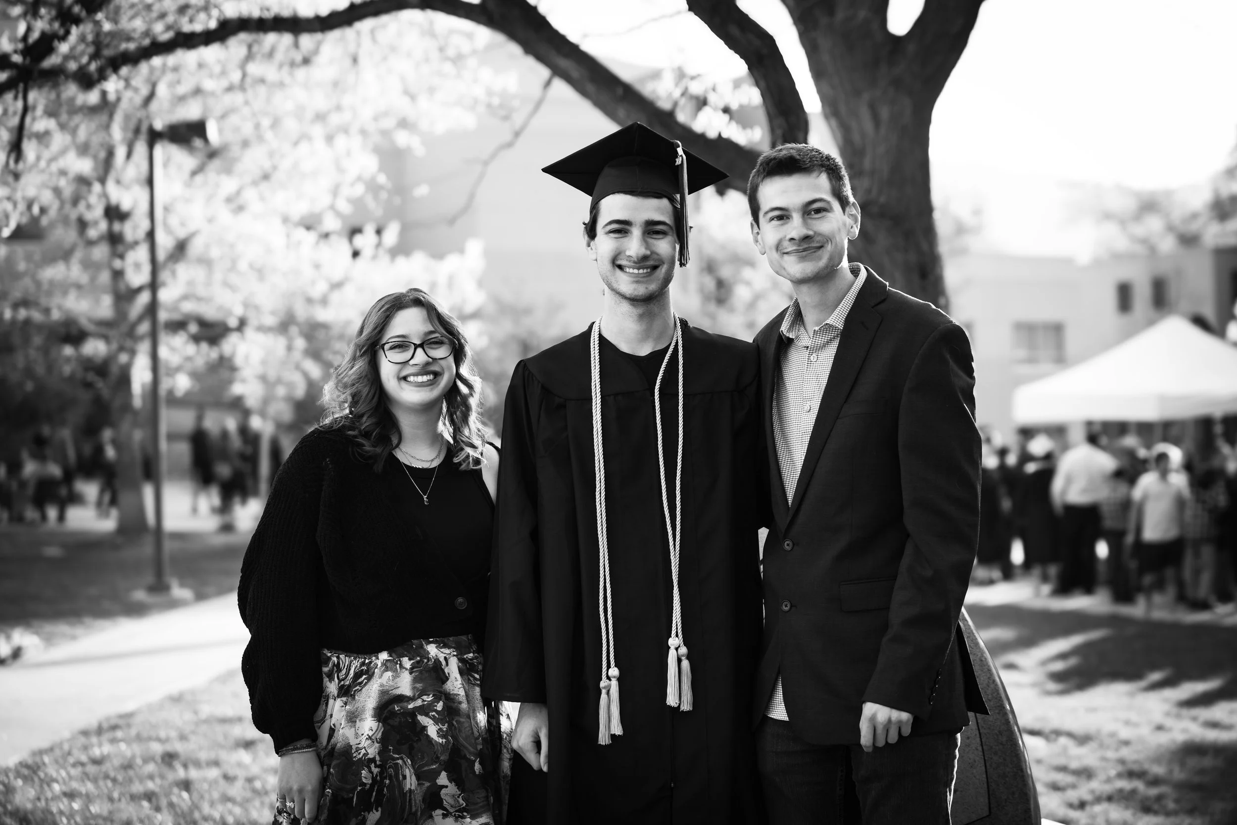 Three young adults outdoors, celebrating graduation. One person wears cap and gown, the others in formal attire, smiling.