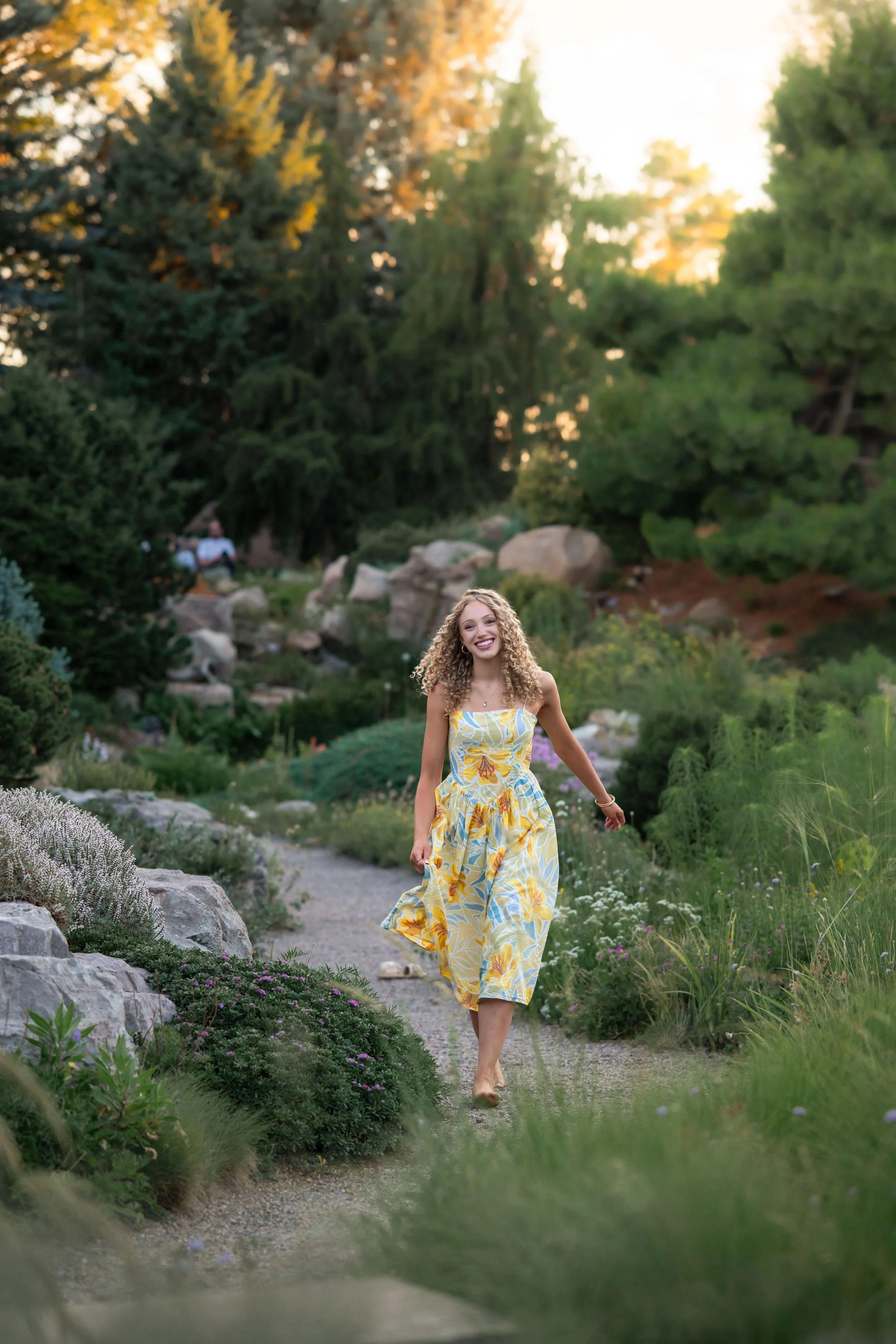 High school senior with curly blonde hair in floral dress with lillies walks through the botanic gardens for her senior pictures.