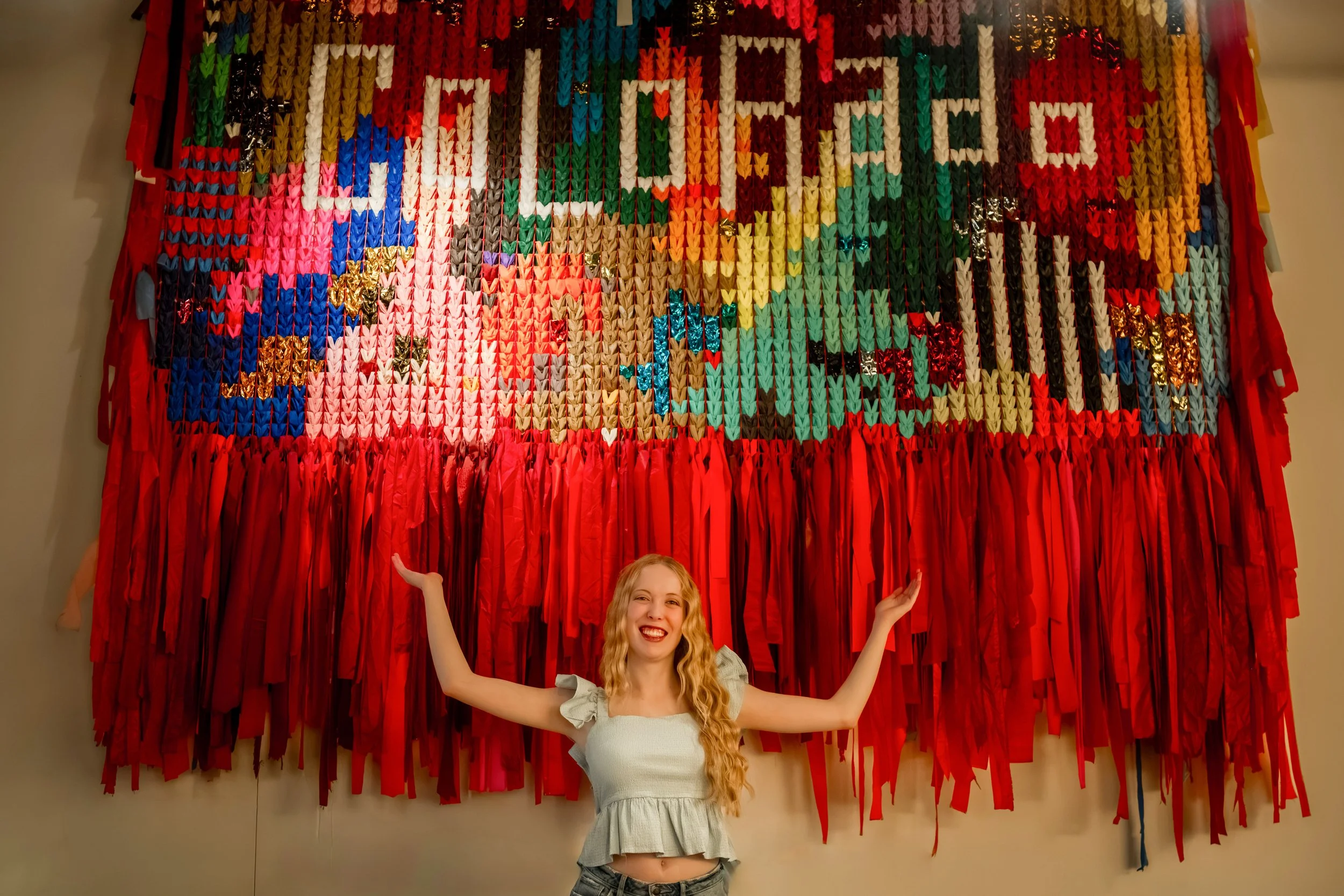 A girl with long blonde wavy hair stands in front of a Colorado tapestry at the Dairy Block in Downtown Denver for her senior photoshoot.