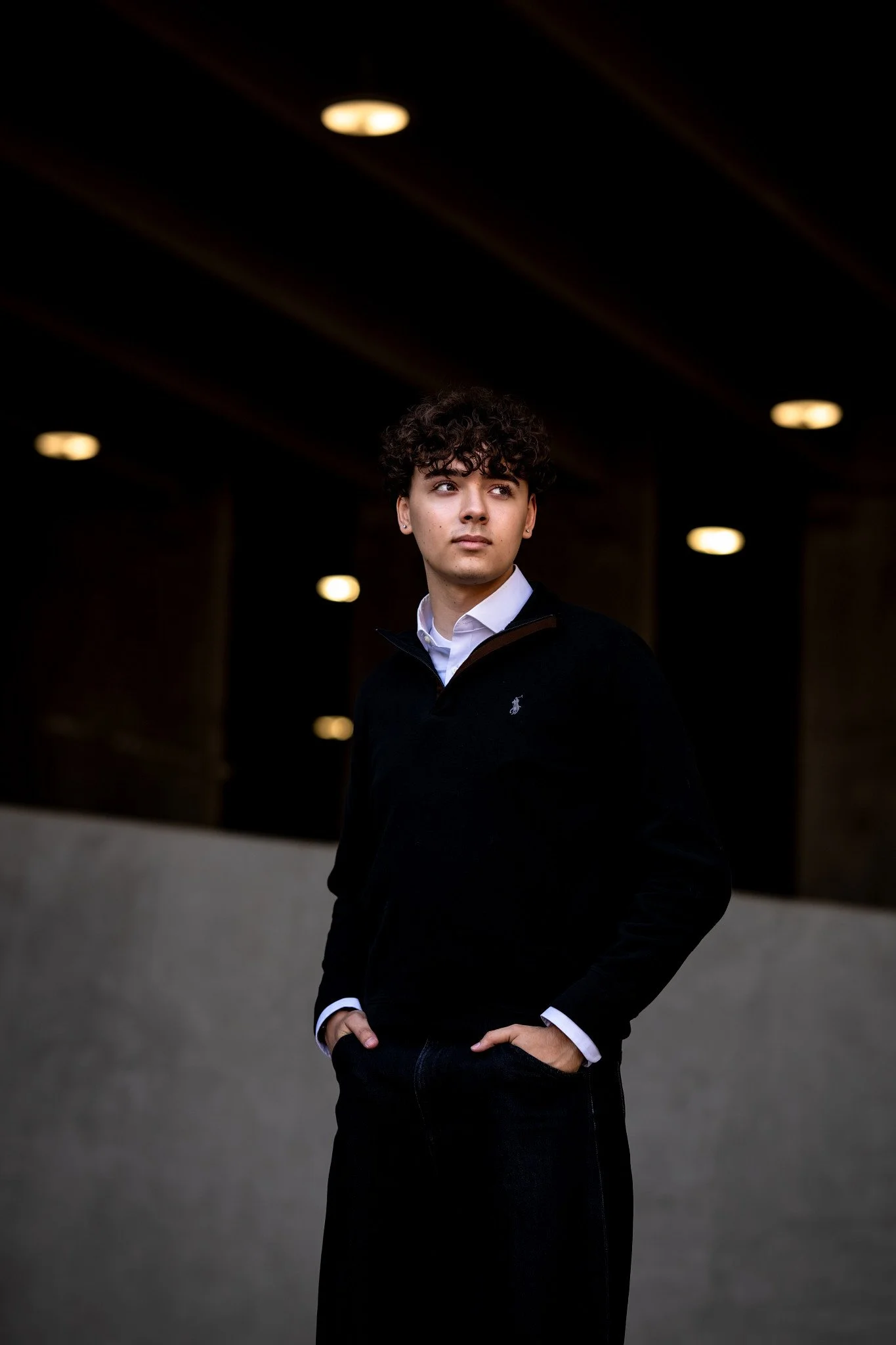 High School Senior boy in a V-neck Polo sweater and dark jeans in a garage looking off into the distance. 
