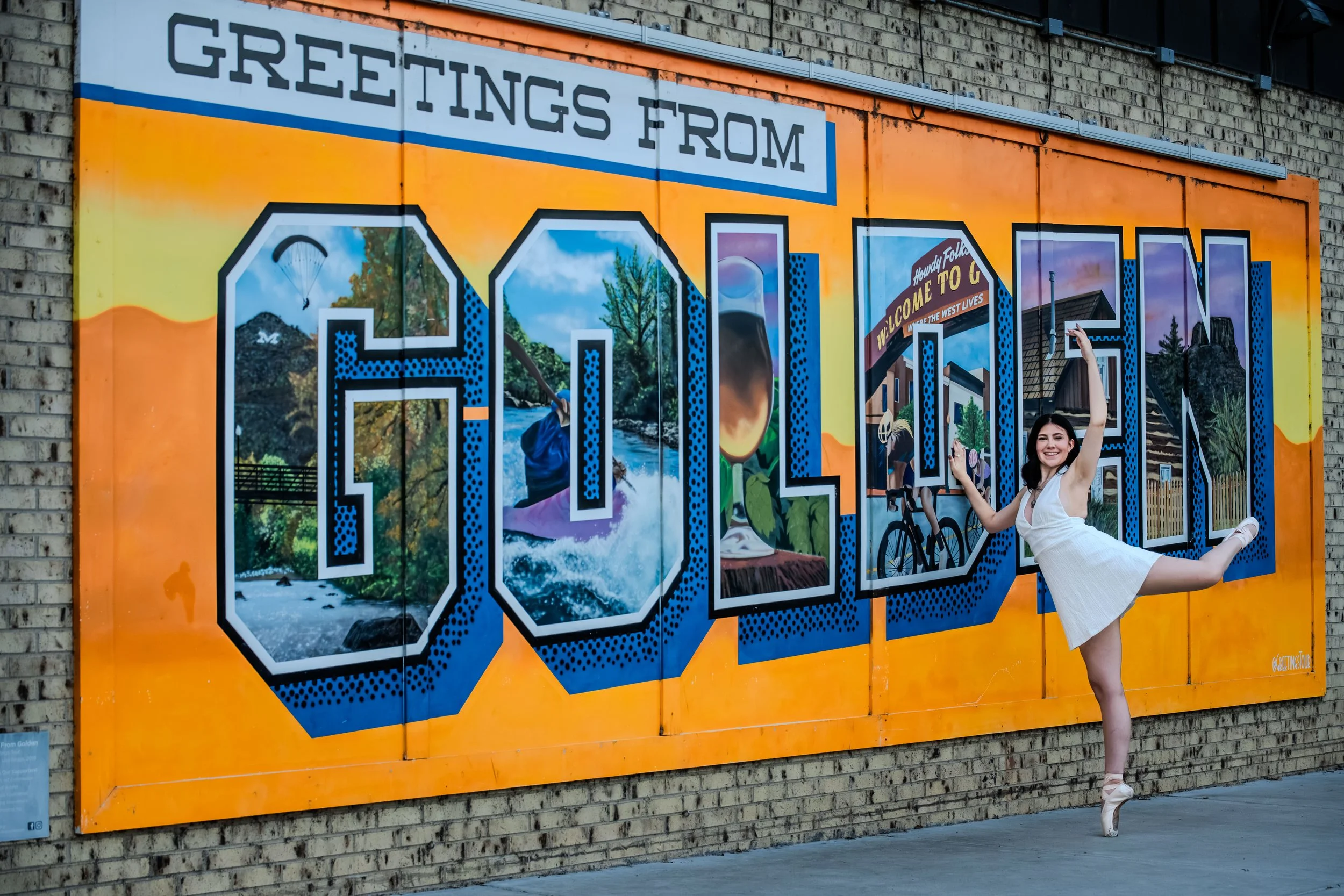 A girl with dark hair wearing a white dress and pointe shoes does a dance move in front of the Golden mural.
