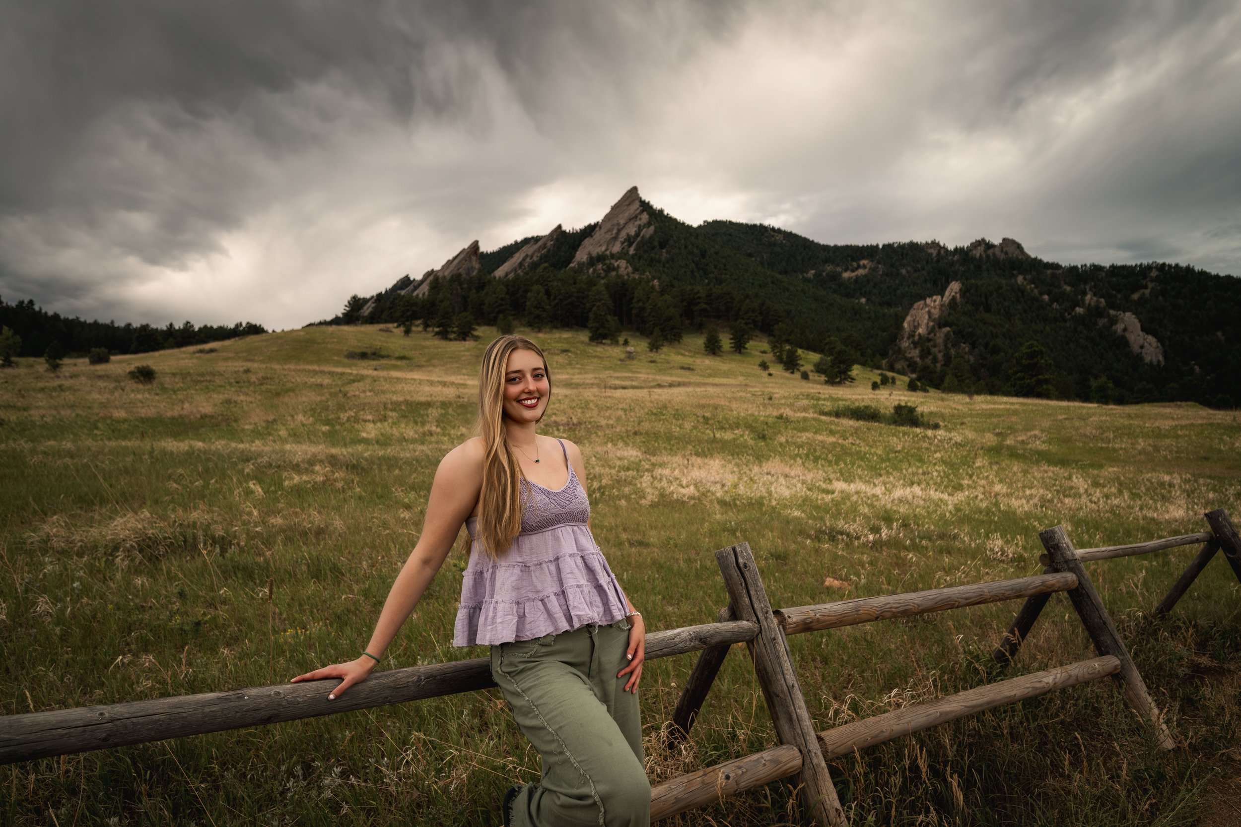 A blonde teen with long hair, wearing a purple tank top and green pants, stands in front of Chautauqua park in Boulder for her senior pictures.