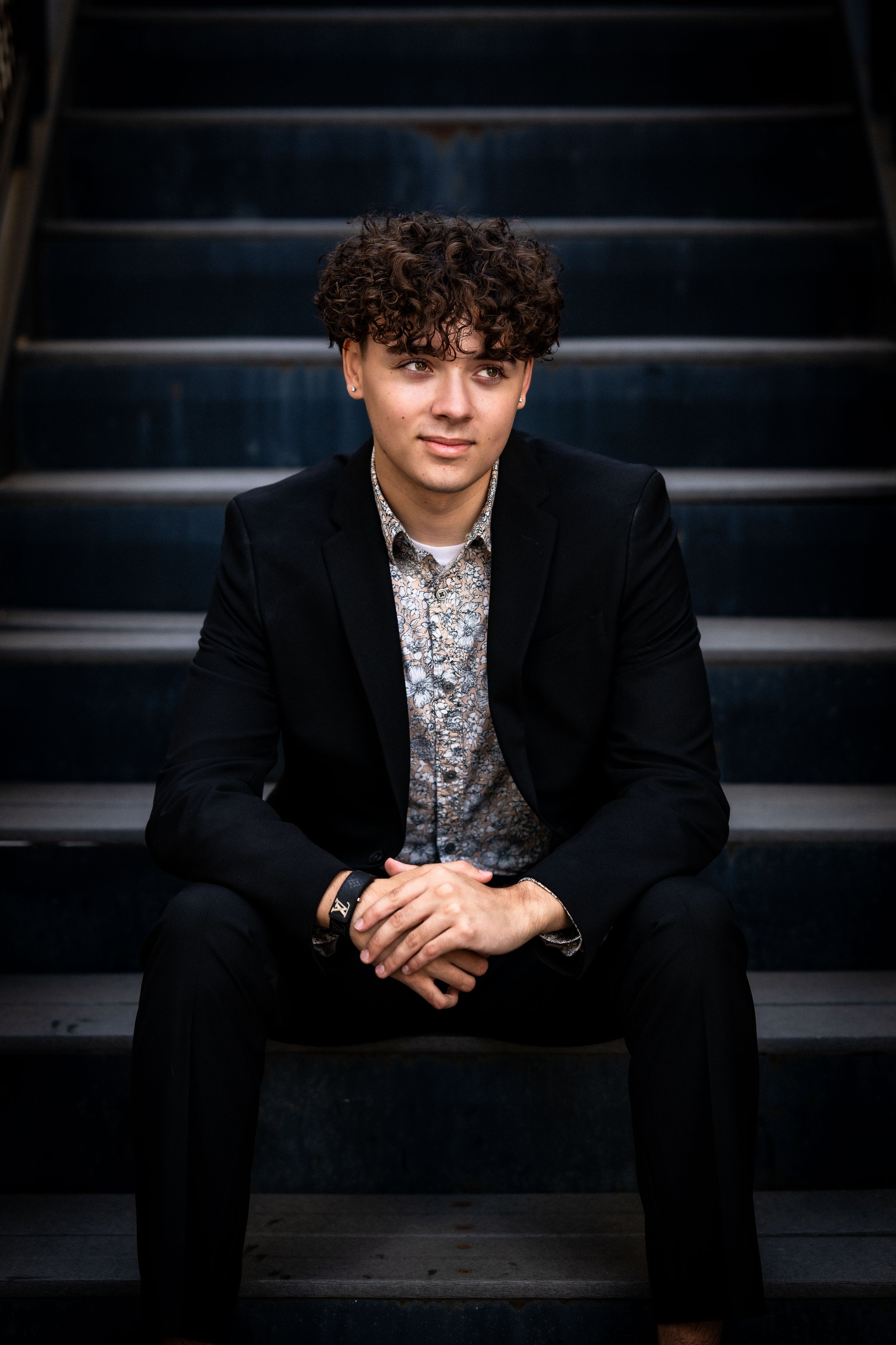 Young man with curly hair, wearing a dark blazer and patterned shirt, sitting on dark stairs, looking to his right.