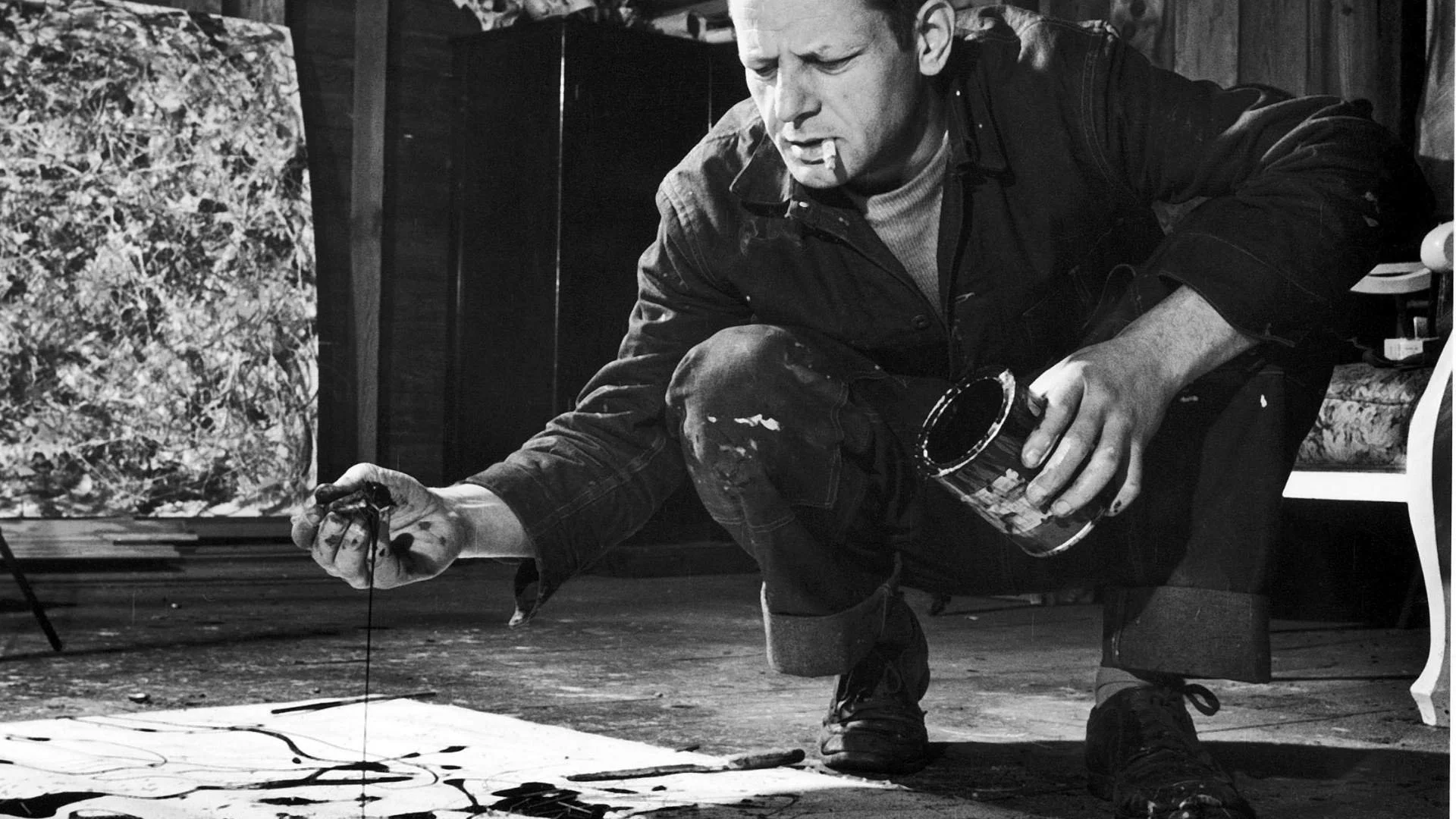 A black and white photograph of the expressionist painter Jackson Pollock at work using his drip technique