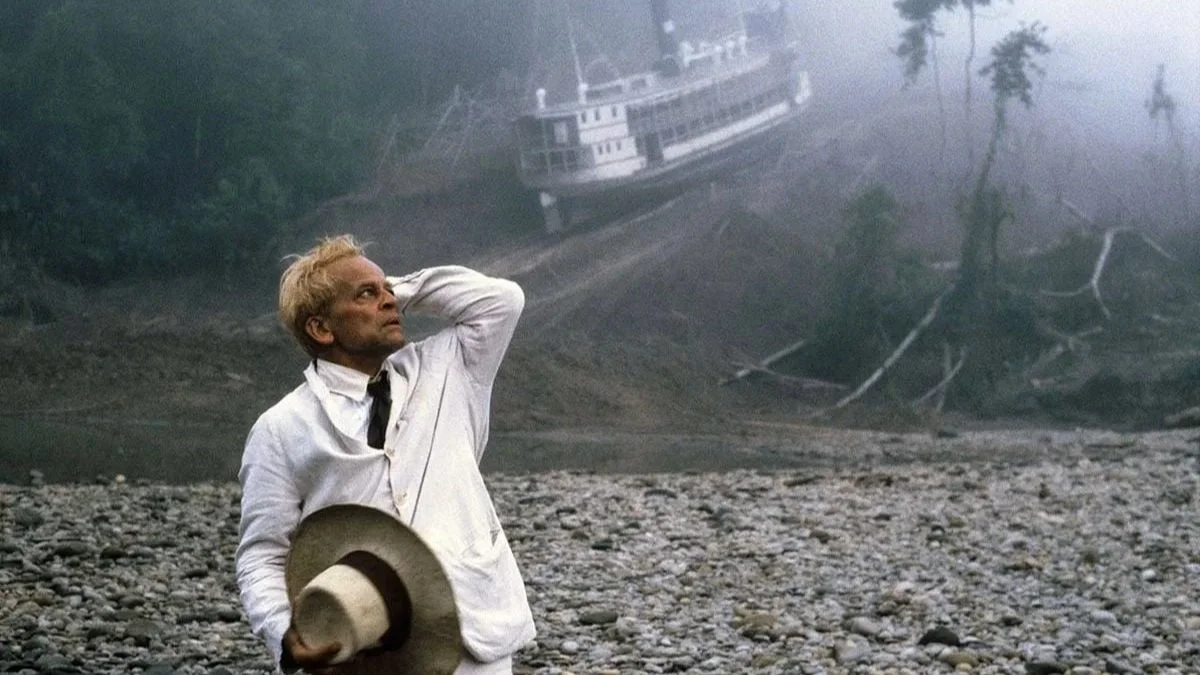 A man in a white suit in the jungle with a boat behind him