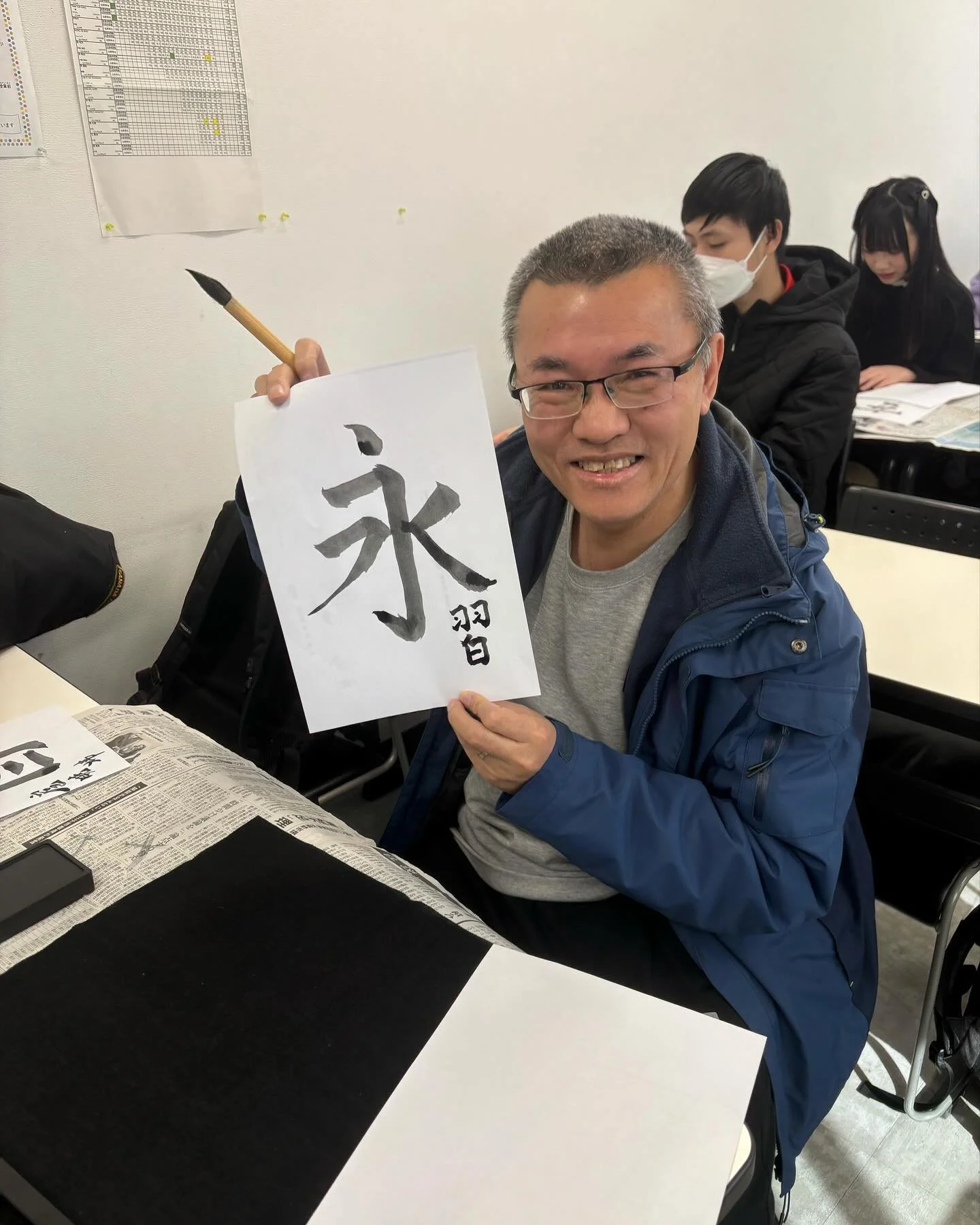 A man smiling and holding up a piece of paper with Japanese calligraphy, sitting at a table covered with newspaper, with other people in the background in a classroom setting.