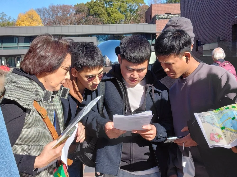 Group of five people, four men and one woman, gathered outdoors looking at papers and a map, with a modern building and trees in the background.