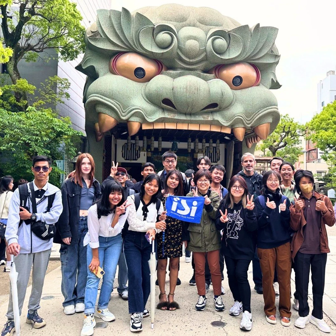 Group of young people smiling and posing for a photo in front of a large Chinese lion head sculpture at a cultural site, with some making peace signs and holding a blue flag.