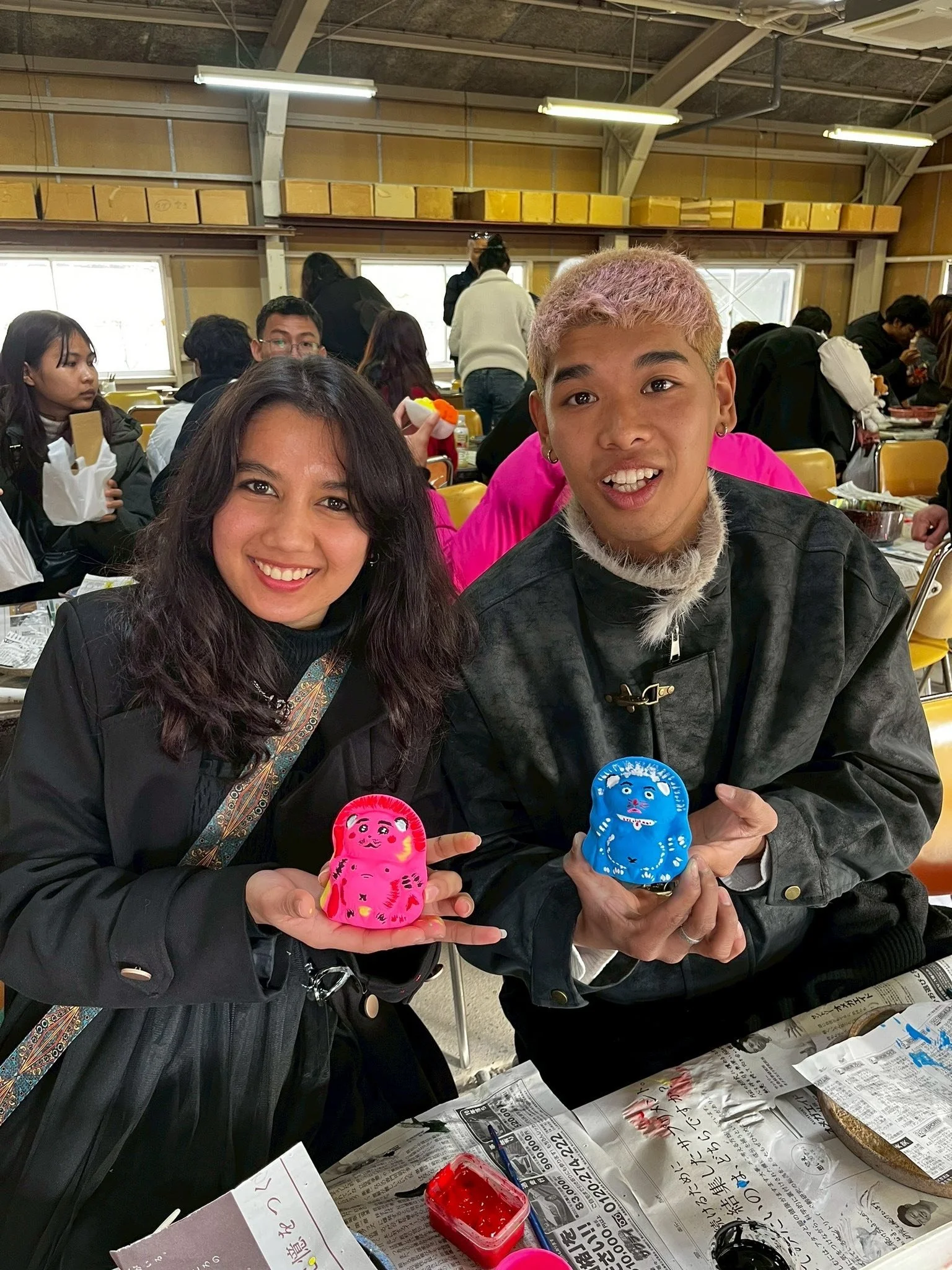Two young people, a woman and a man, sitting at a table with newspapers, holding painted figurines in the shape of traditional Japanese dolls. The woman has dark hair and is smiling, the man has light-colored hair and is smiling slightly. The background shows a room with other people engaged in similar activities, and the ceiling has exposed beams and fluorescent lighting.