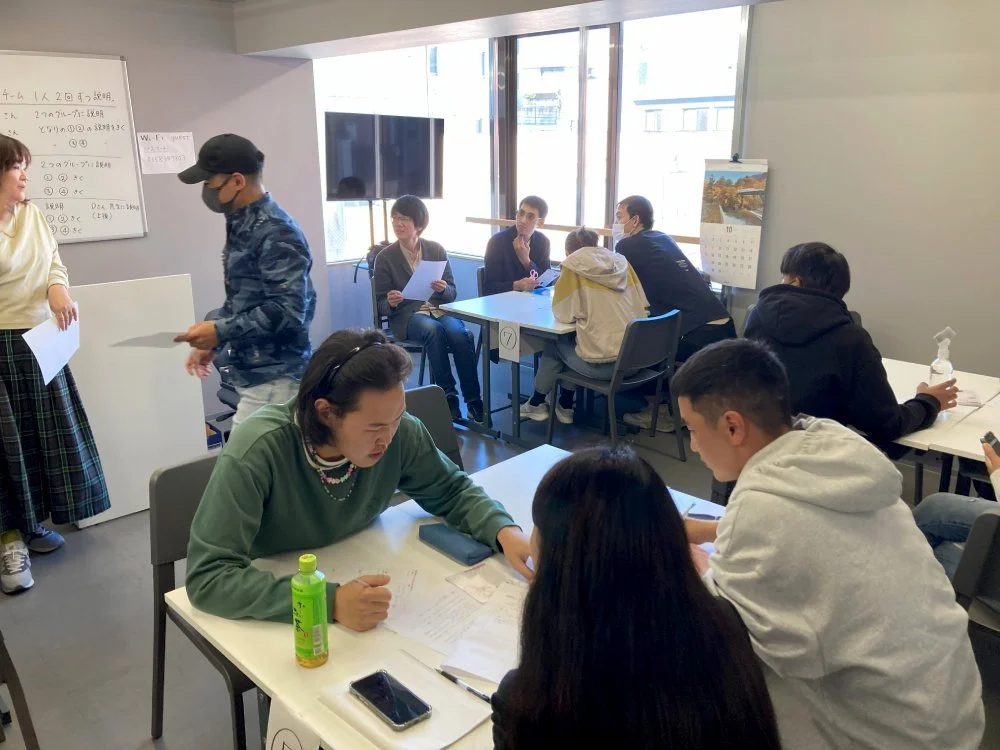 A group of students and teachers in a classroom engaged in discussions and activities, with some students sitting at tables and a whiteboard on the wall.