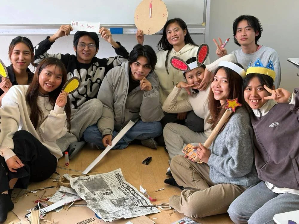 Group of nine diverse young people sitting on the floor in a classroom, some holding paper props like yellow ear shapes and bunny ears, with a clock and whiteboard behind them. They are smiling and making peace signs.