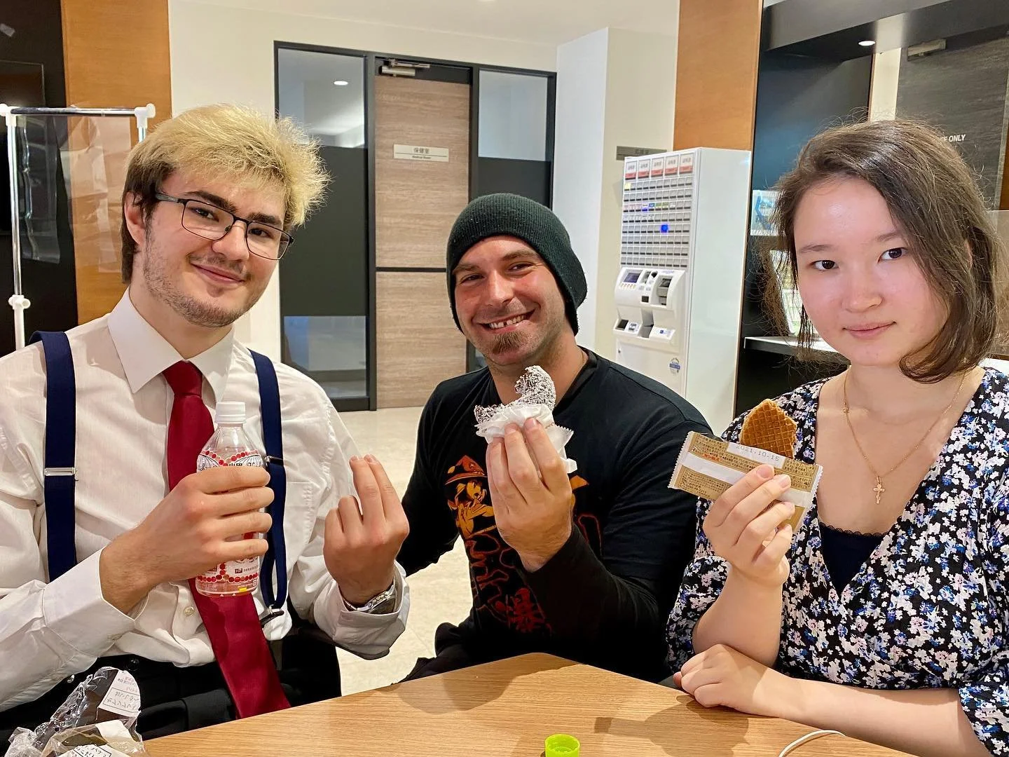 Three people sitting at a table in a building, holding snacks and drinks, smiling at the camera. The man on the left is wearing glasses, a white shirt, a red tie, and suspenders, holding a water bottle. The man in the middle is wearing a black beanie and a black shirt, holding a foil-wrapped snack. The woman on the right is wearing a patterned top and holding a snack bar, with a cross necklace around her neck.