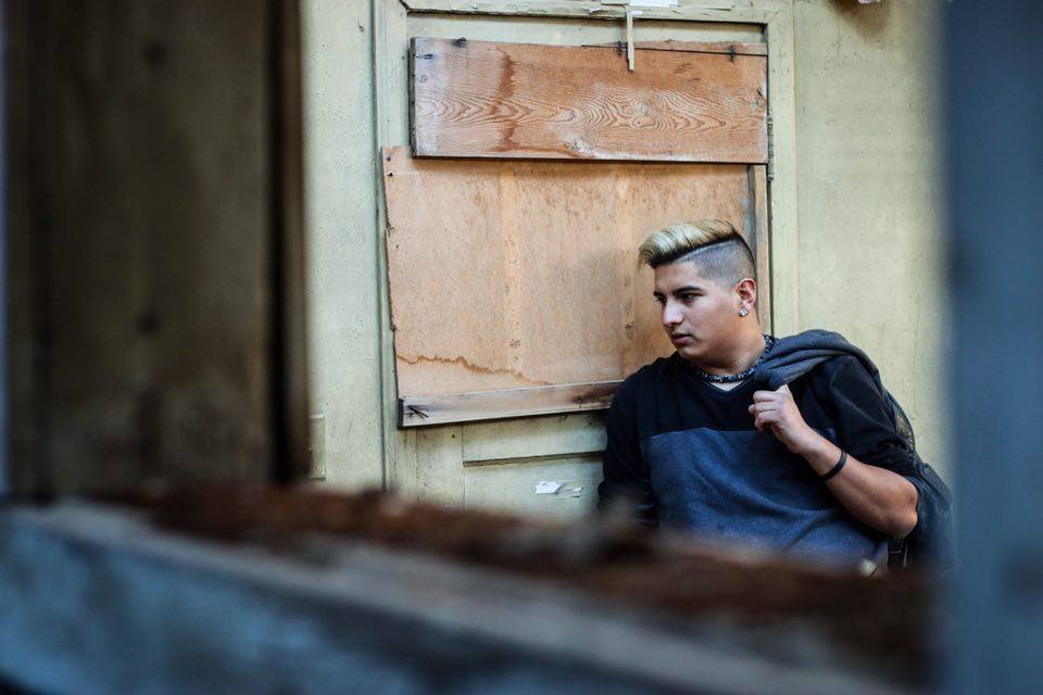 A young man with a blonde mohawk hairstyle sitting against a weathered wooden wall with boarded-up window, wearing a dark hoodie, holding a strap of backpack, looking to the side in a contemplative manner.
