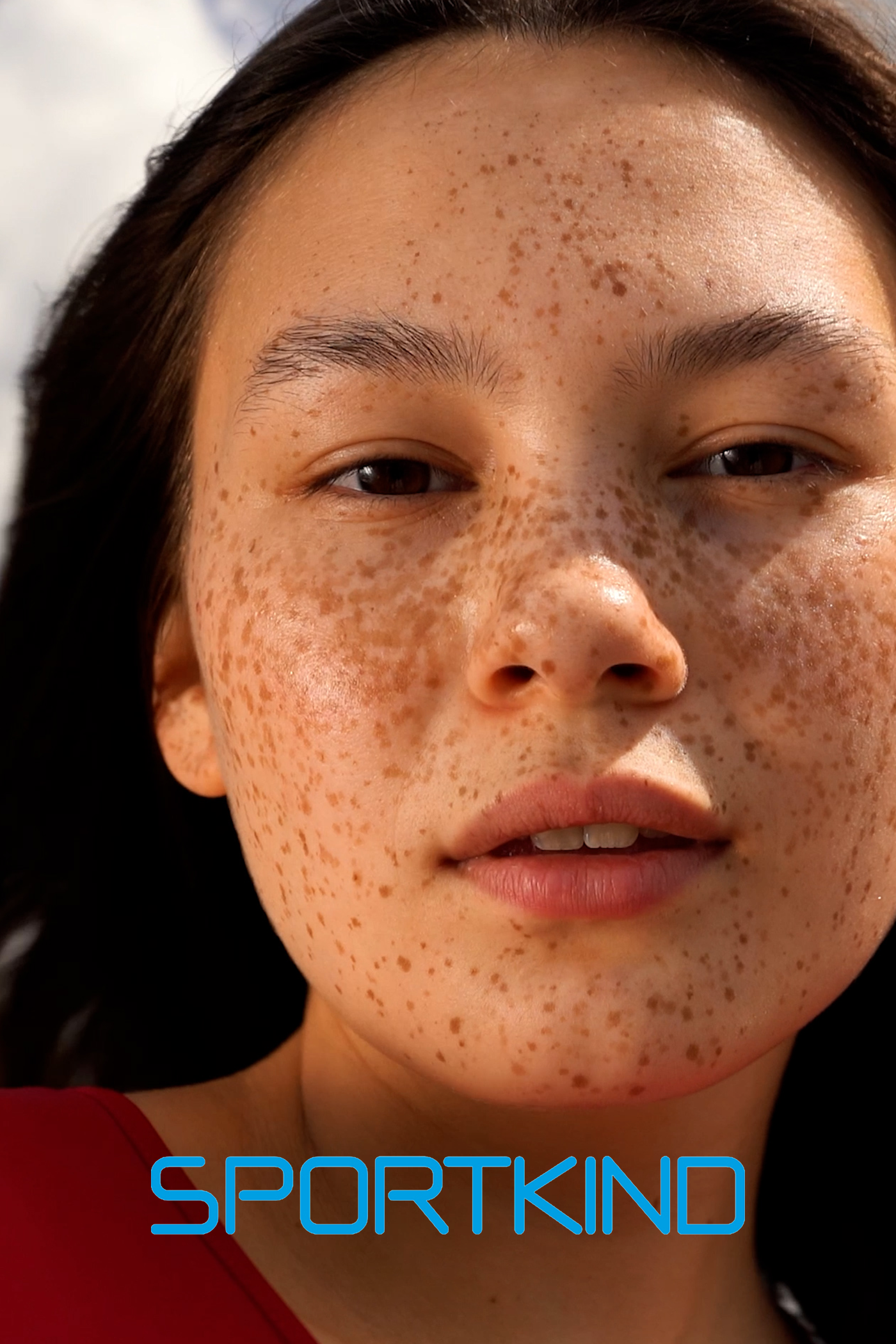 Close-up of a woman's face with freckles and dark hair, wearing a red top, with the word 'SPORTKIND' written in blue text at the bottom. - Commercial Client of Kantara Productions