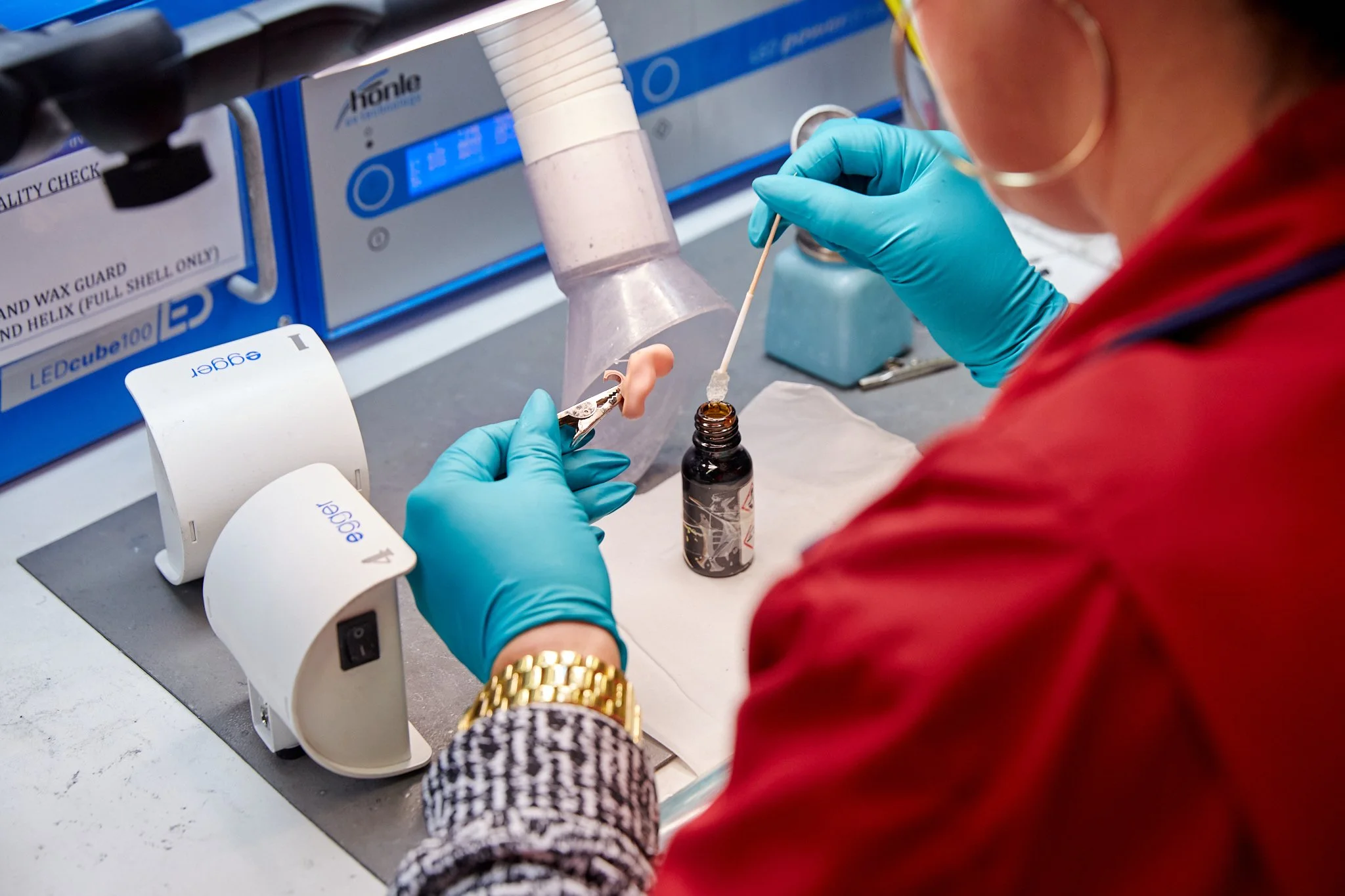 A person with a gold watch and patterned clothing is working with a lab technician in blue gloves to perform a scientific procedure. The setting appears to be a laboratory with equipment, including a test tube and a vaporizer or extraction device.