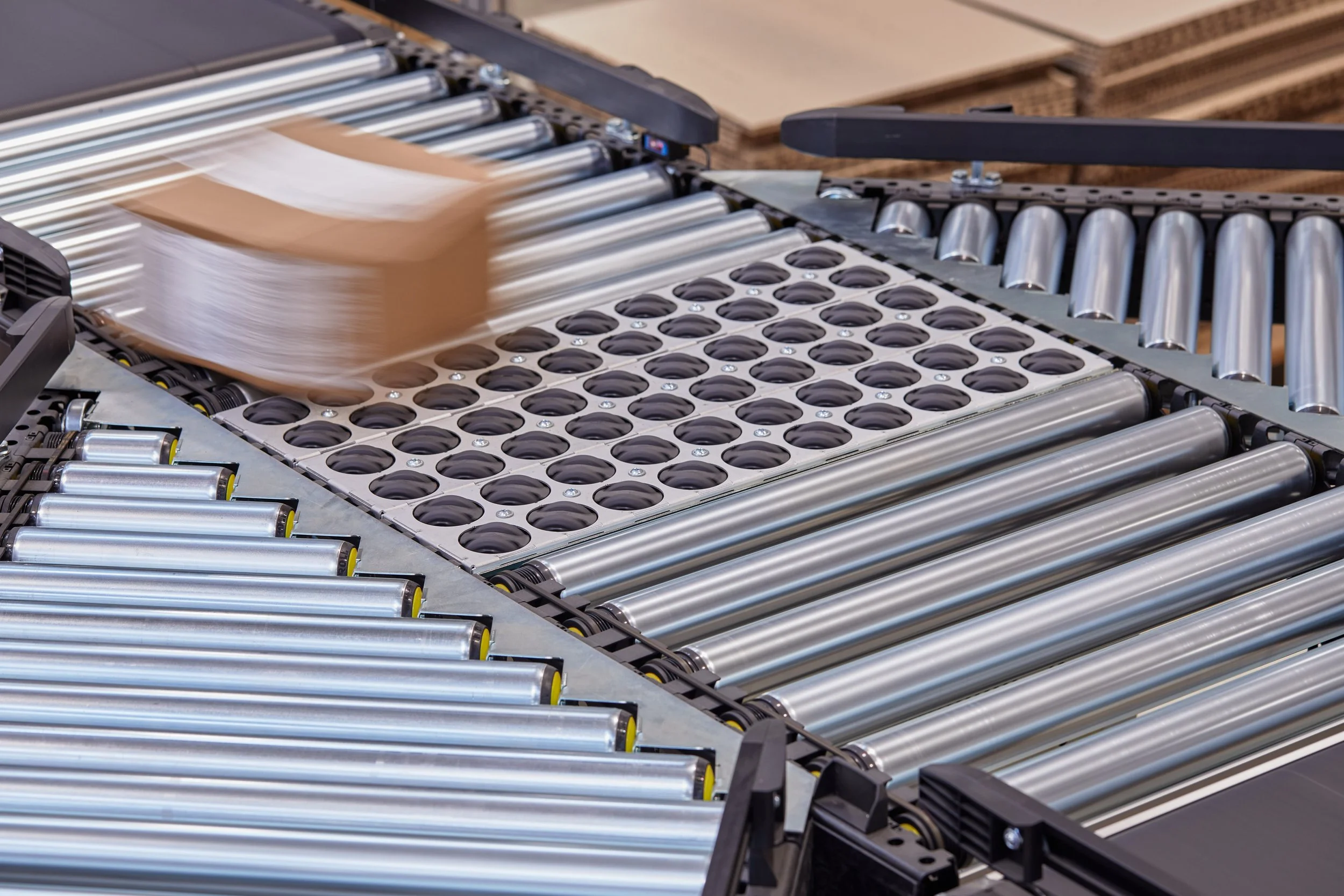 Conveyor belt with multiple rollers moving a cardboard box with white paper attached on top.