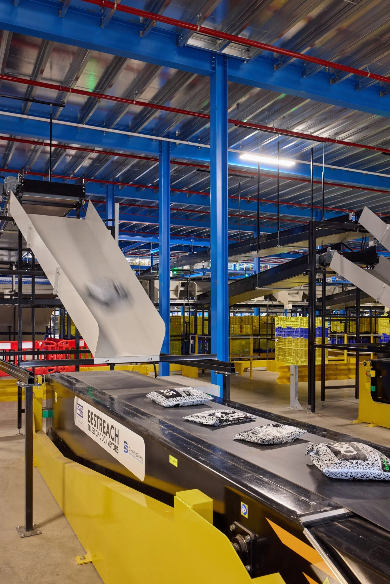 Inside a warehouse with blue steel shelving and a conveyor system. The conveyor is black and yellow, with three packages on it. The packages are wrapped in black and white patterned paper. There are stacked yellow plastic bins in the background.