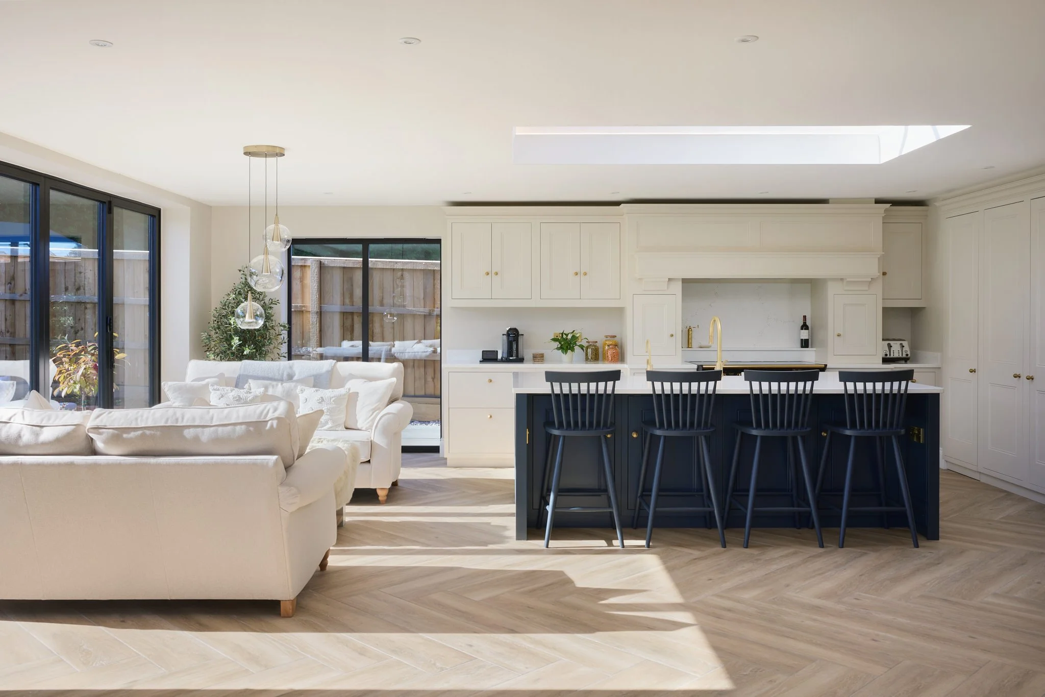 Modern kitchen and living room with cream-colored sofa, white cabinetry, black bar stools, large sliding glass doors, and a skylight in the ceiling.
