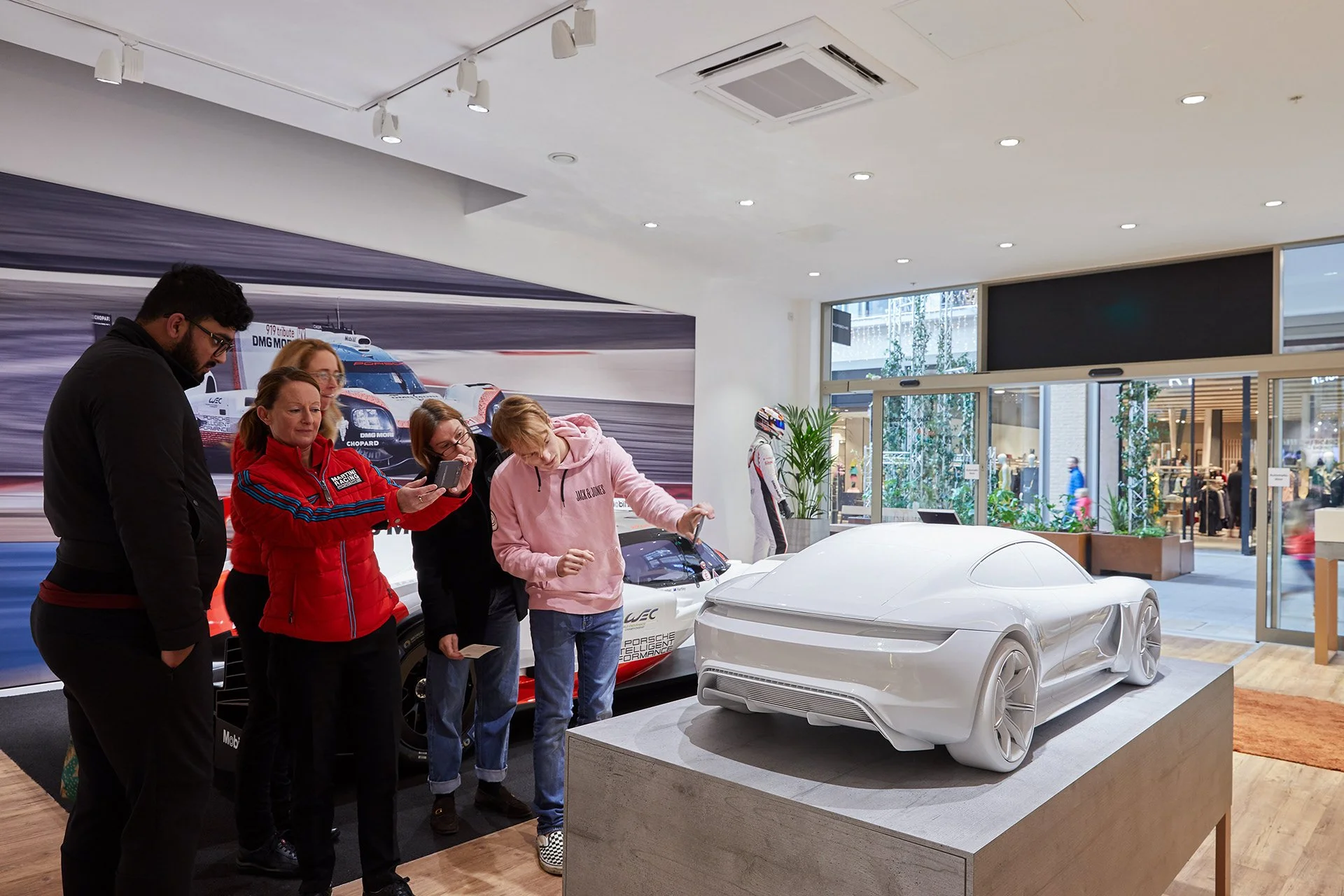 Interior Retail Photography a group of five people, four women and one man, looking at an indoor display of a futuristic white car model with smooth curves and aerodynamic design, in a showroom with a large racing-themed mural on the wall