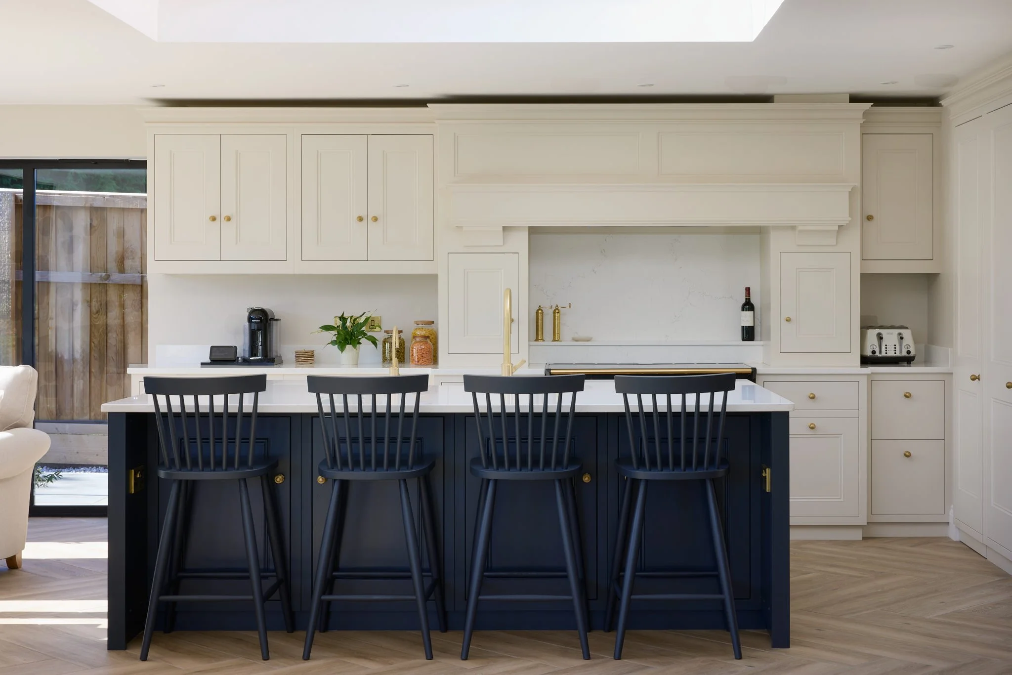 Modern kitchen with white cabinetry, marble backsplash, and white countertops. A navy blue island with black chairs is in the foreground. Items on the counter include a coffee maker, plant, jars, a wine bottle, and toaster. Sliding glass door with ve