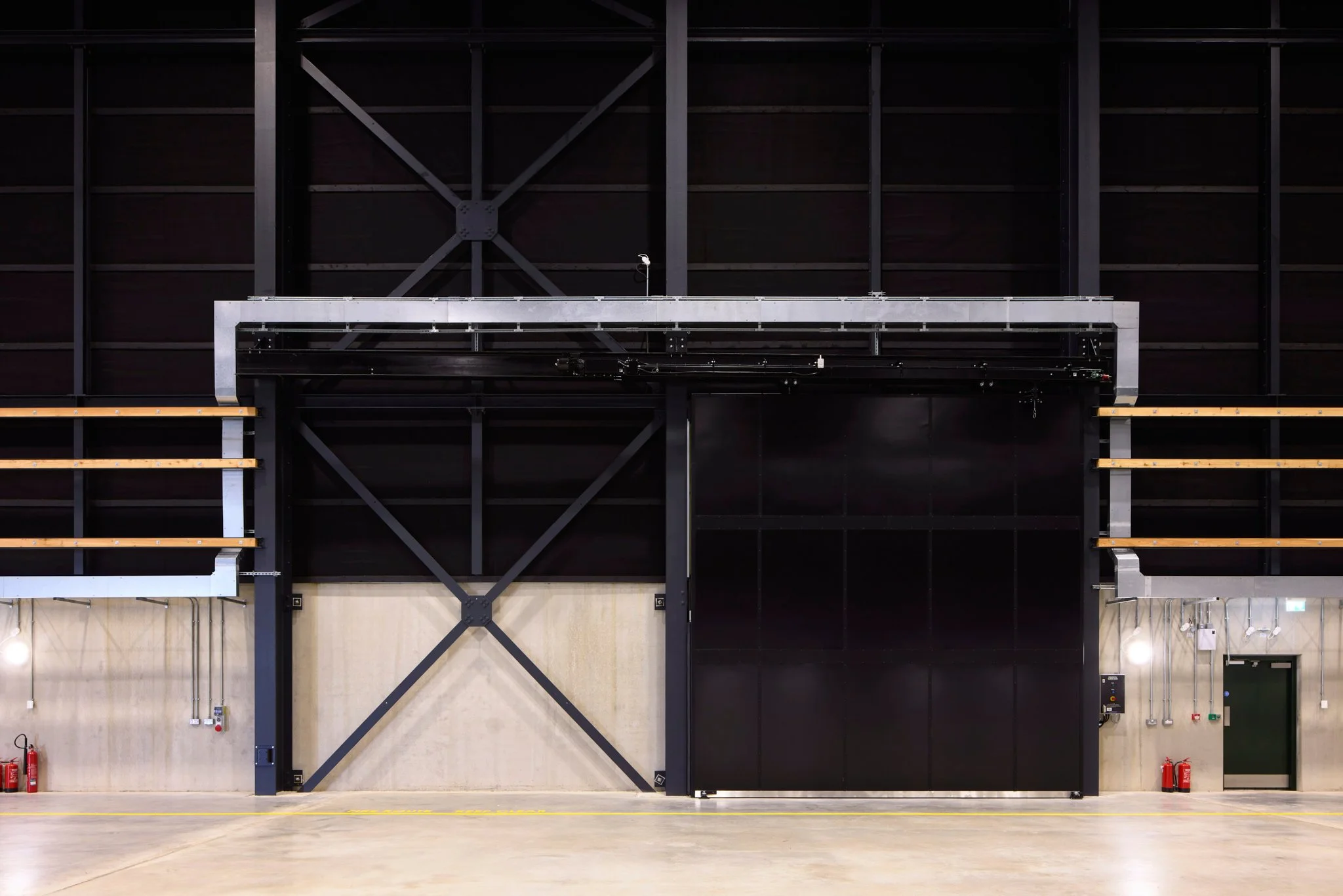 Interior of an industrial building with a large, black sliding door, metal framework, wooden stairs, fire extinguishers, and industrial control panels.
