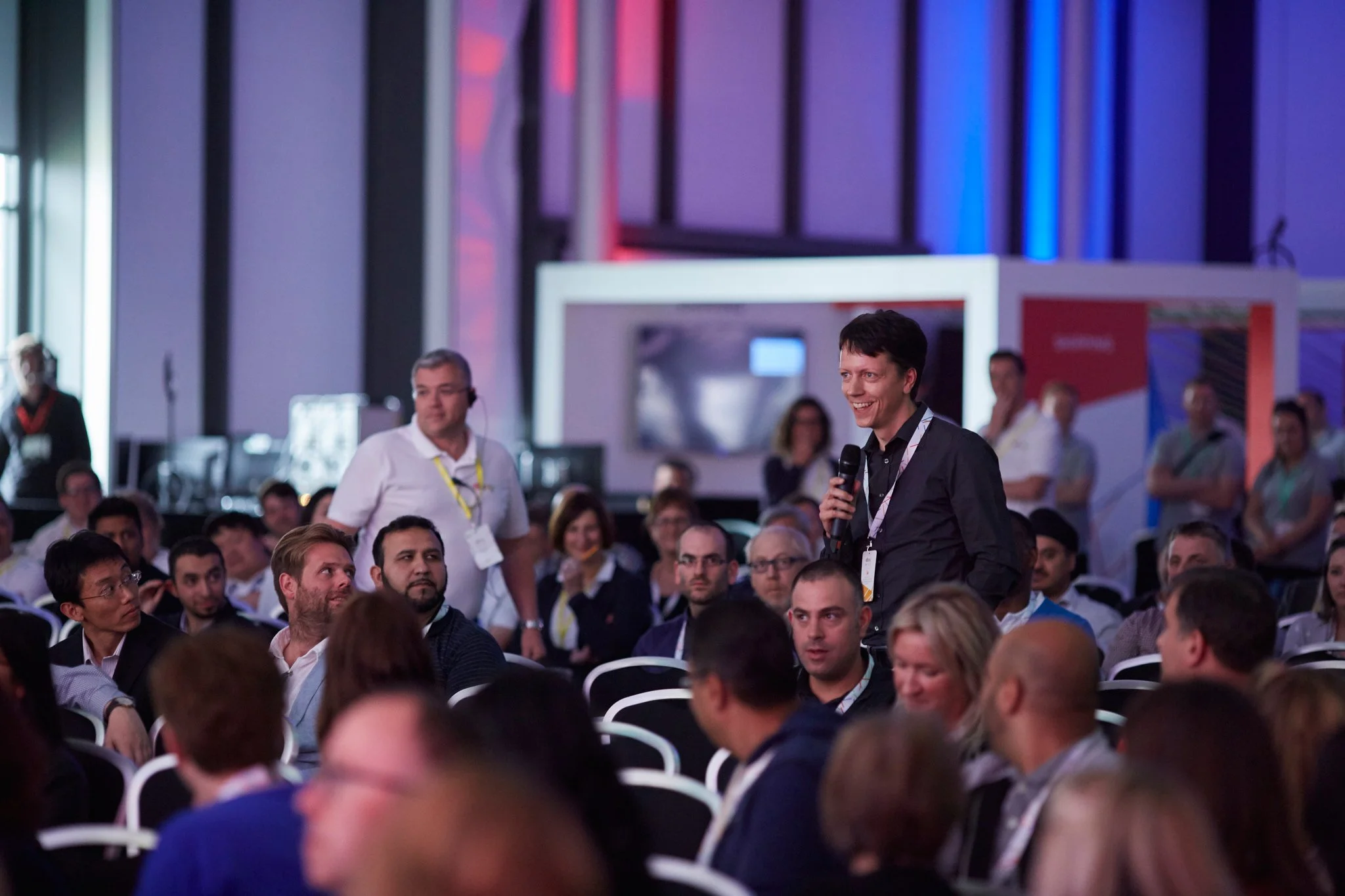 A man is speaking into a microphone during a conference or seminar, with an audience listening attentively, in a large, modern indoor venue with vibrant lighting.