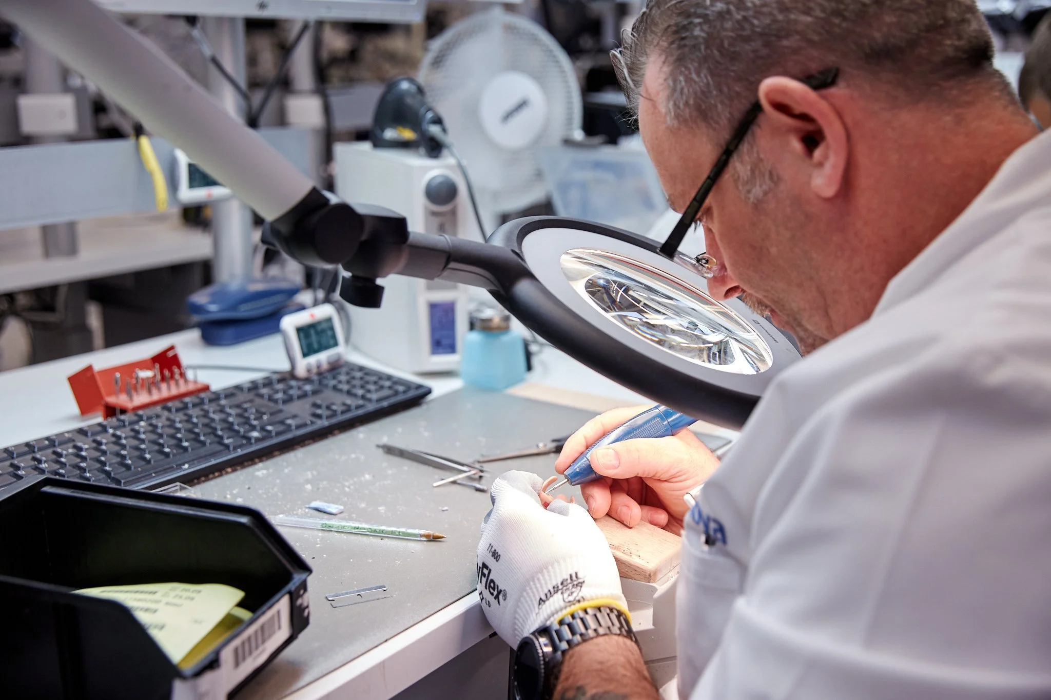 A scientist in a lab coat and safety glasses examines a small object with a tool, surrounded by various laboratory equipment like a microscope, tools, and electronic devices.