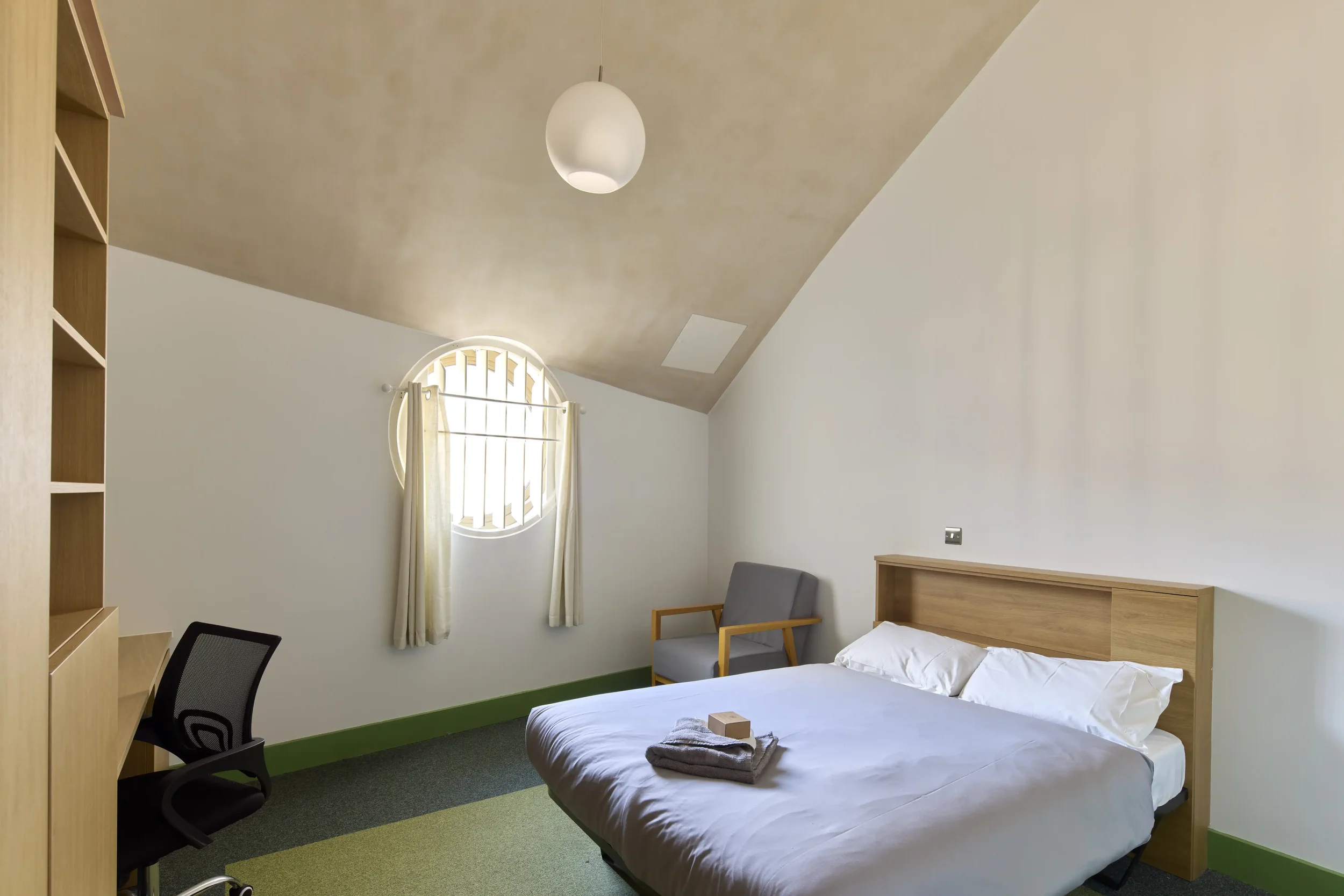 A minimalistic hotel room with a bed, a gray armchair, a wooden desk, a black office chair, and a window with white curtains. The room has white walls, a green carpet, and a sloped beige ceiling with a ceiling light.