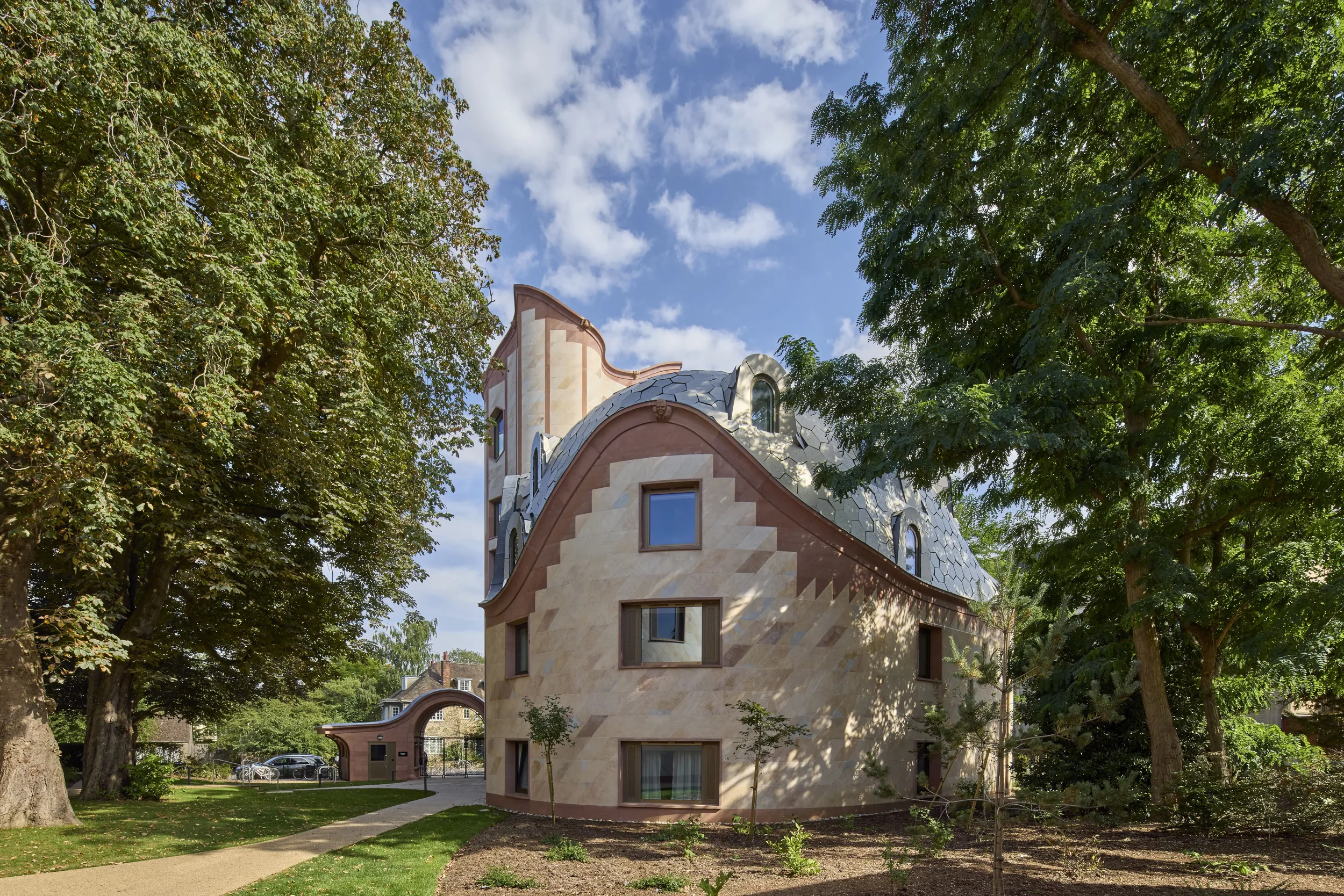 A unique house with an artistically curved roof and window designs, surrounded by green trees and a park, under a partly cloudy sky.