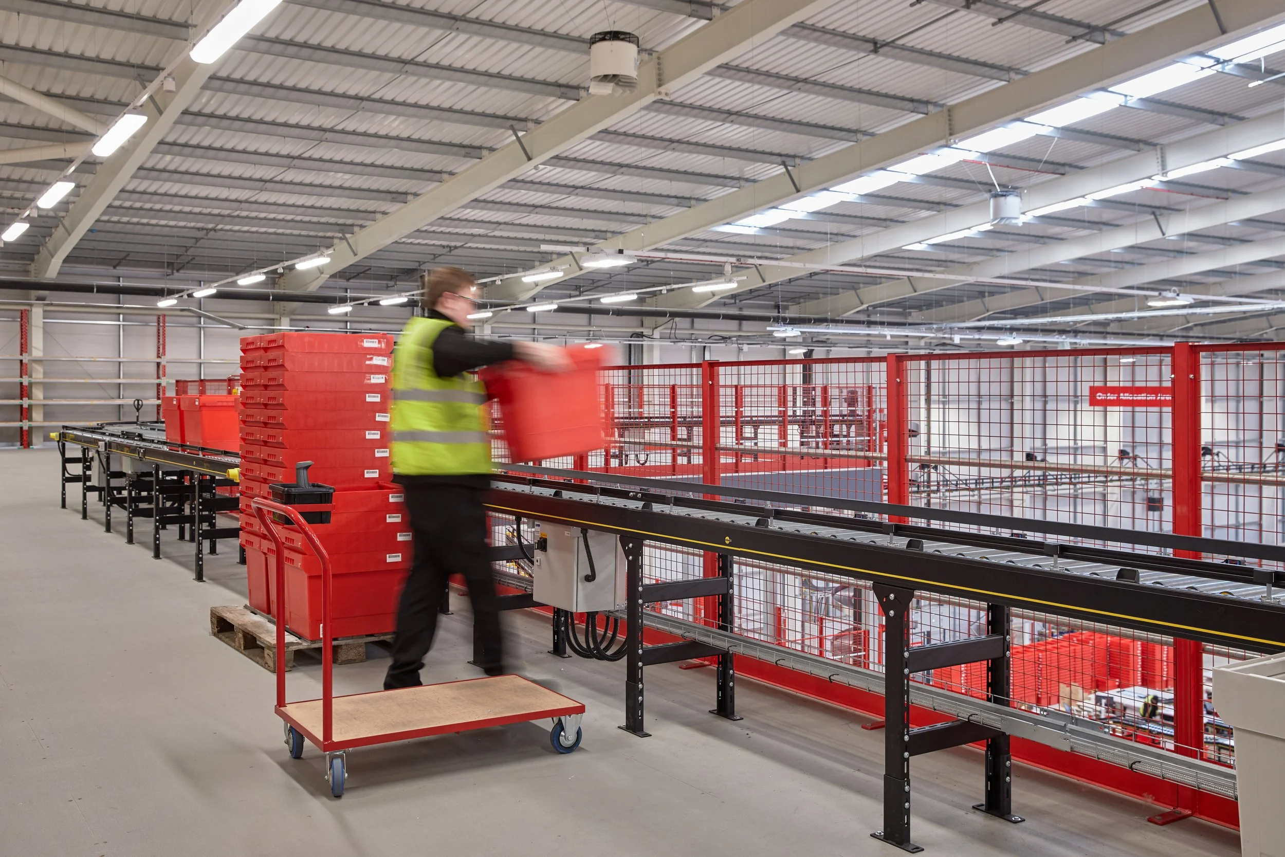 A person wearing a yellow safety vest and black clothing is moving a large red container in a warehouse. The warehouse has high ceilings, bright lighting, metal storage racks, and red safety barriers.