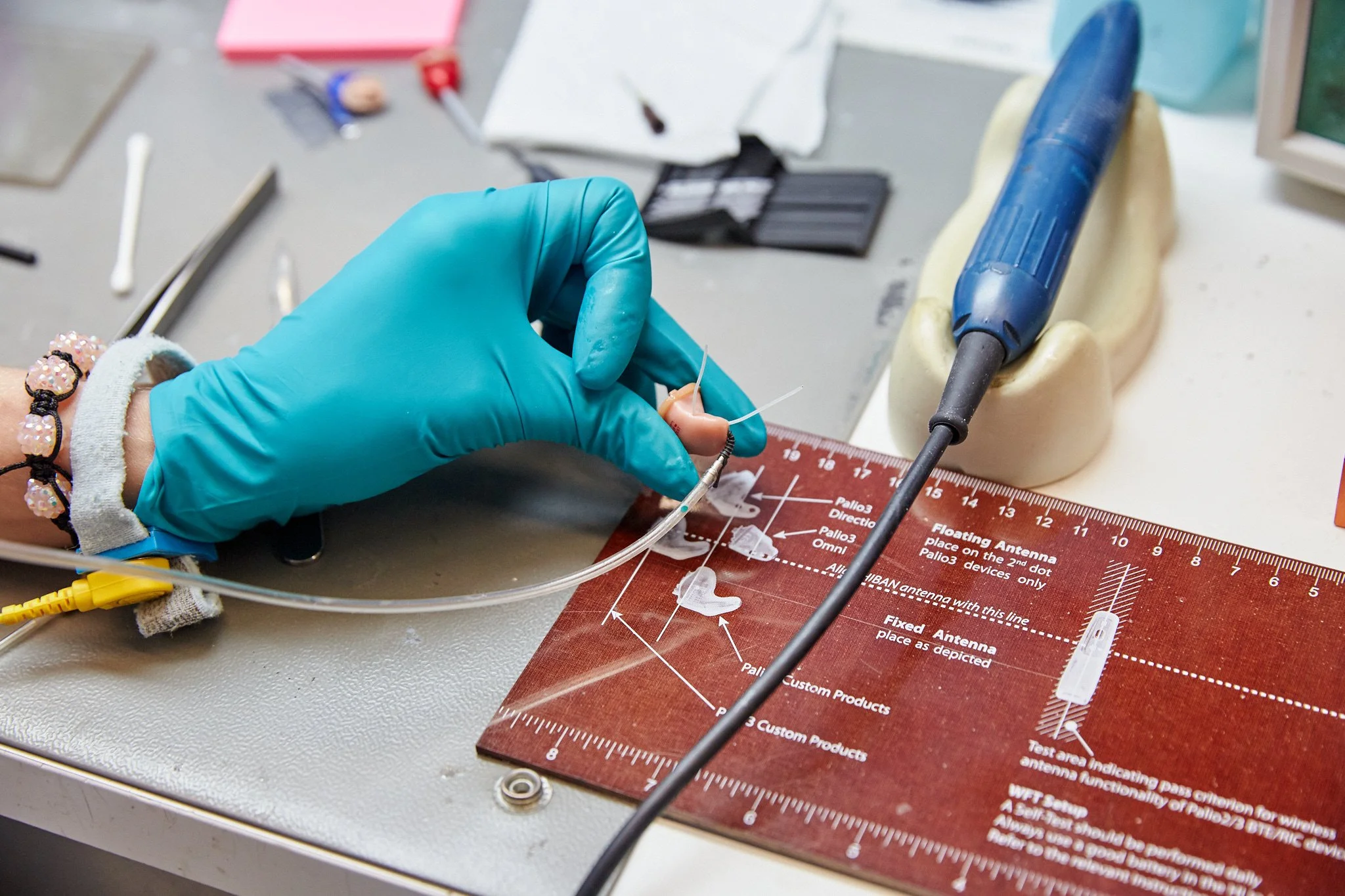 Close-up of a gloved hand working with a small tool on a tiny object, surrounded by various tools and equipment on a work surface engaged in precision work, possibly in a laboratory or workshop setting.