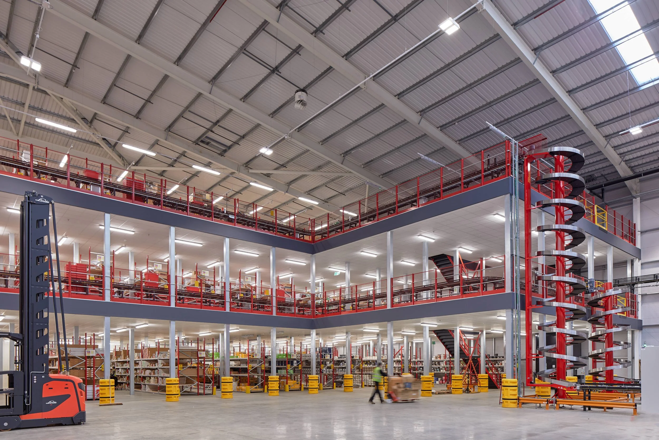 Interior of a large warehouse or store with multiple shelves and a spiral staircase, a worker moving boxes, and industrial lighting.