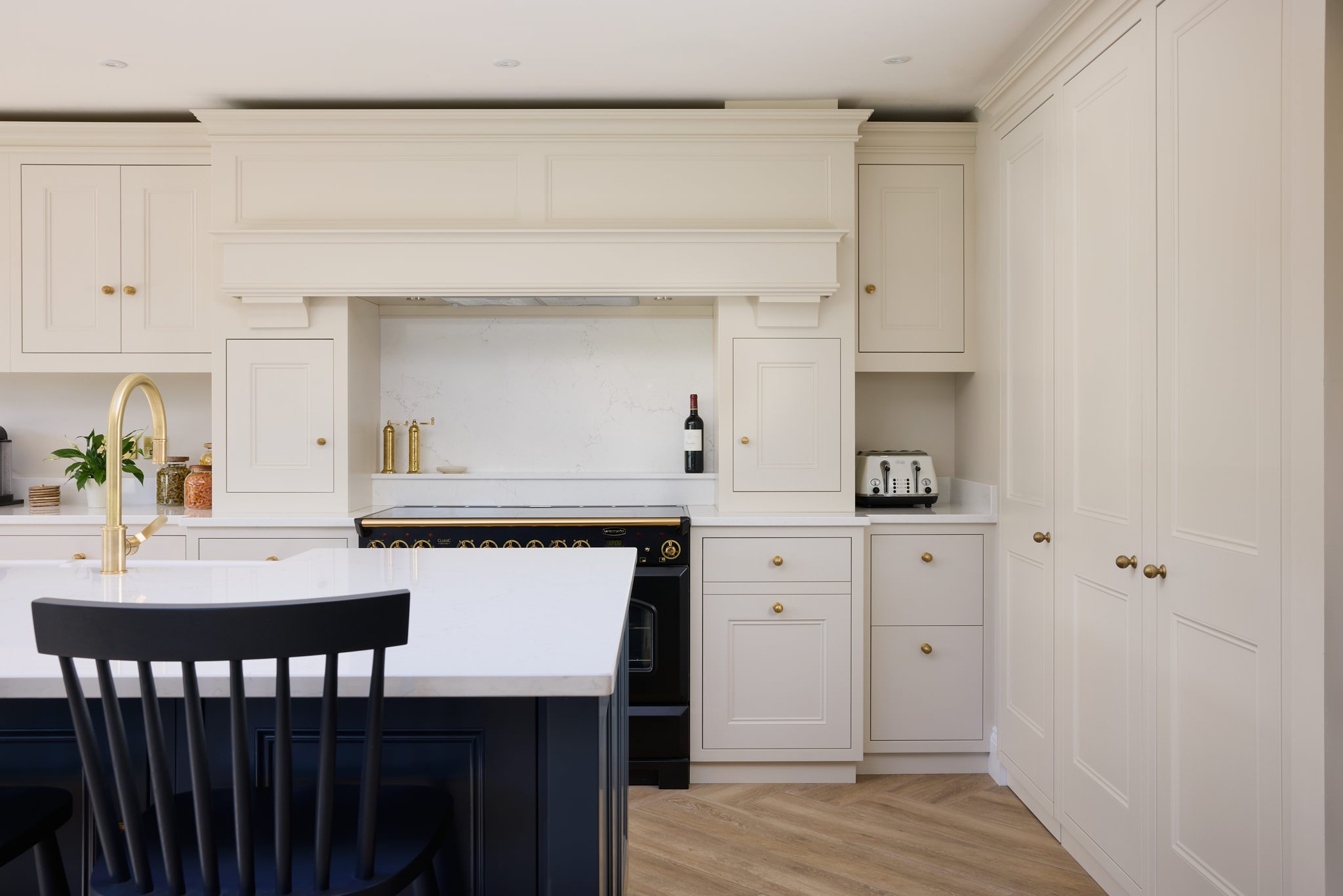 A modern kitchen with white cabinets, a white countertop, and a black island with a white top. There is a gold faucet and gold cabinet handles, with some small appliances including a toaster, and a wine bottle on the back wall. Wooden flooring is vis