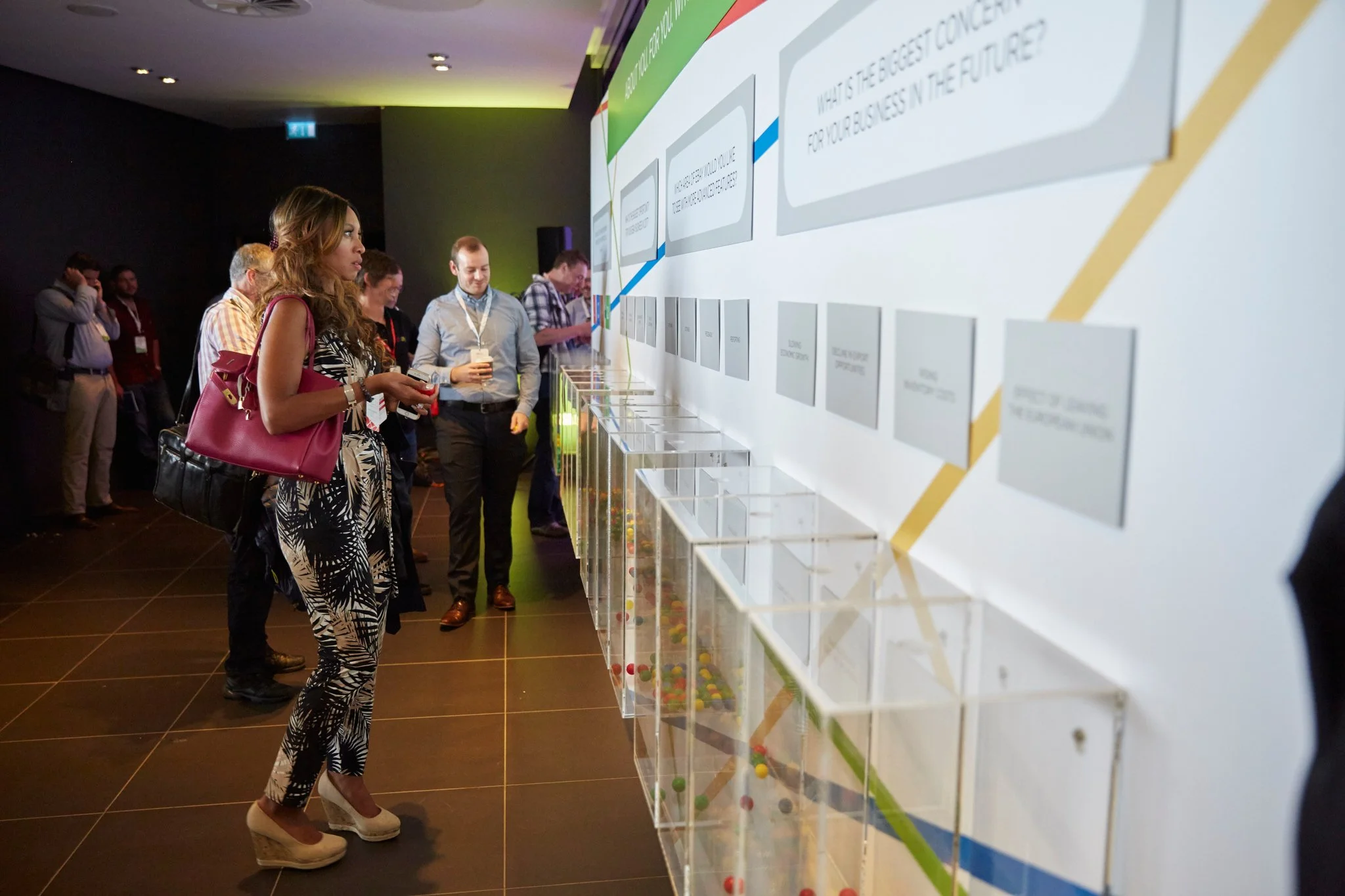 People attending a conference or event, standing in front of an interactive display with questions and transparent boxes filled with color balls, in a dimly lit room with dark walls.