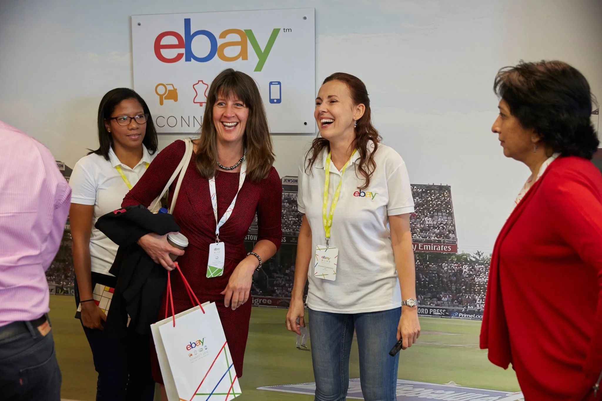 Group of women standing and talking in front of an eBay sign at an event, smiling and laughing.