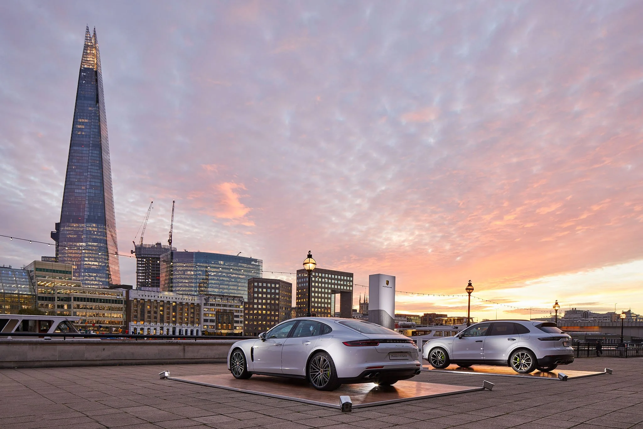 A cityscape at sunset with modern skyscrapers, including the Shard, and several parked white cars in the foreground.