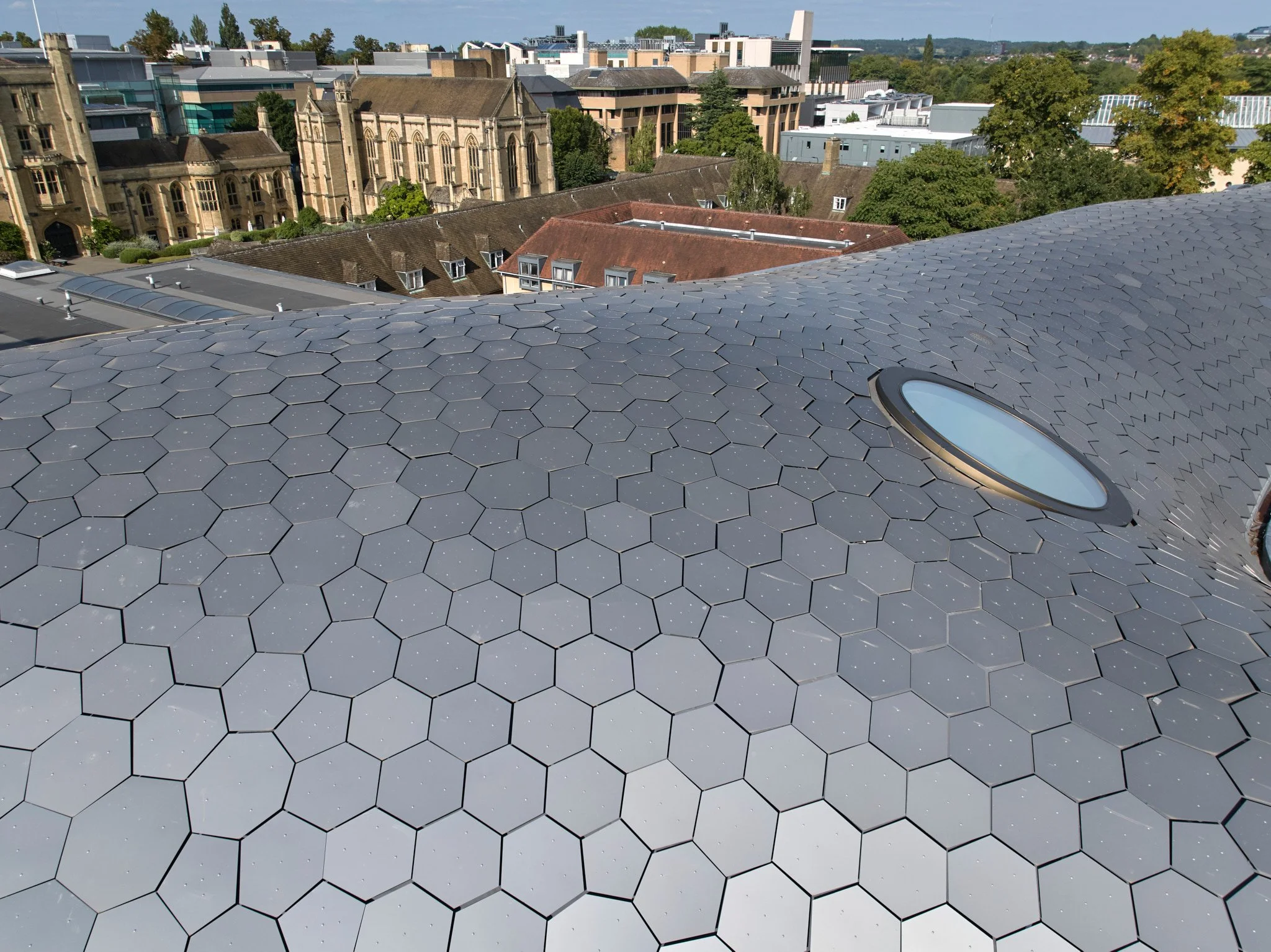 A rooftop covered with hexagonal shingles and a skylight. In the background, city buildings and green trees are visible under a sunny sky.