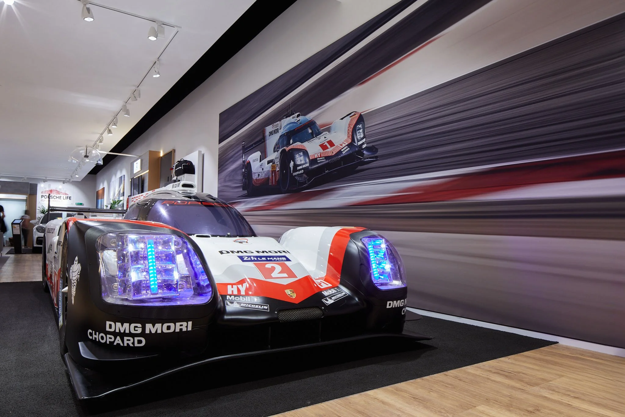 Race cars on display in a showroom with a large photograph of a race car on the wall behind them.