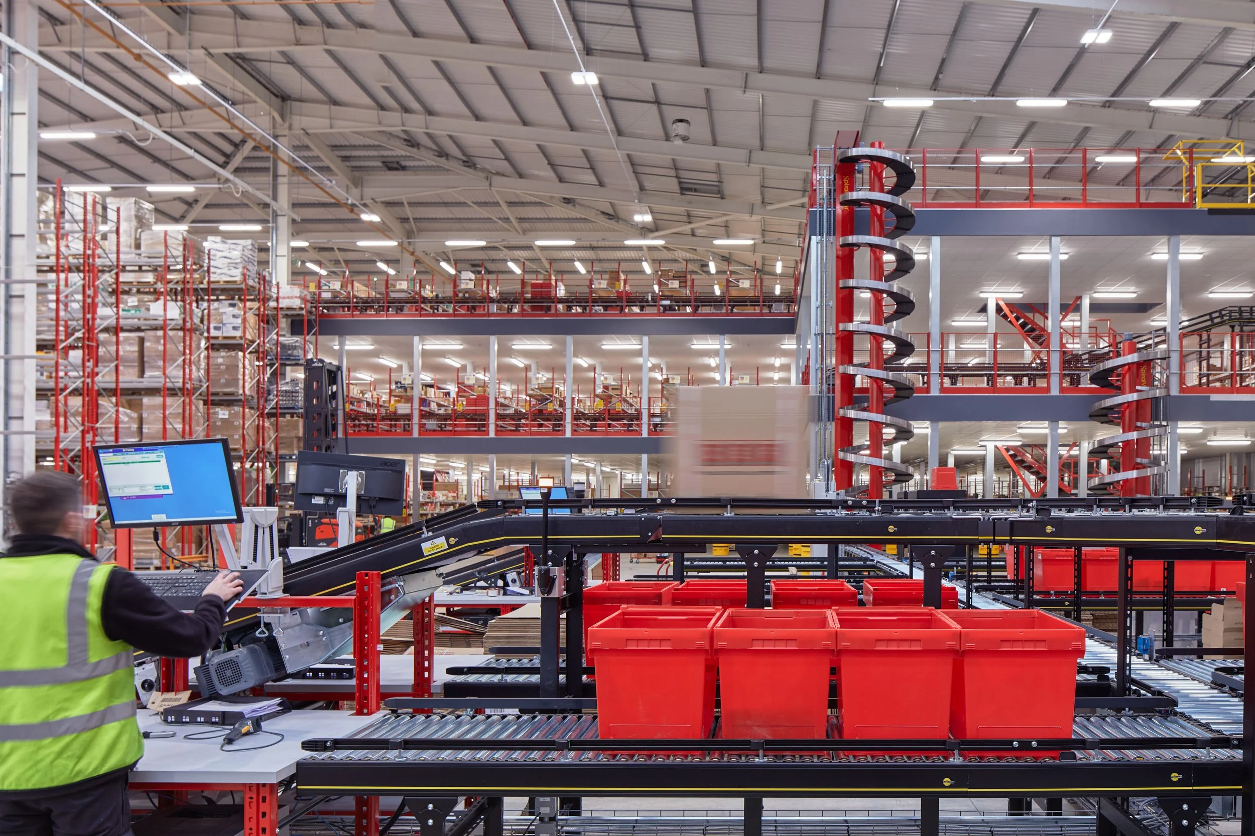 Inside a warehouse with red shelving and conveyor belts, a worker in a yellow safety vest operates a computer at a workstation.