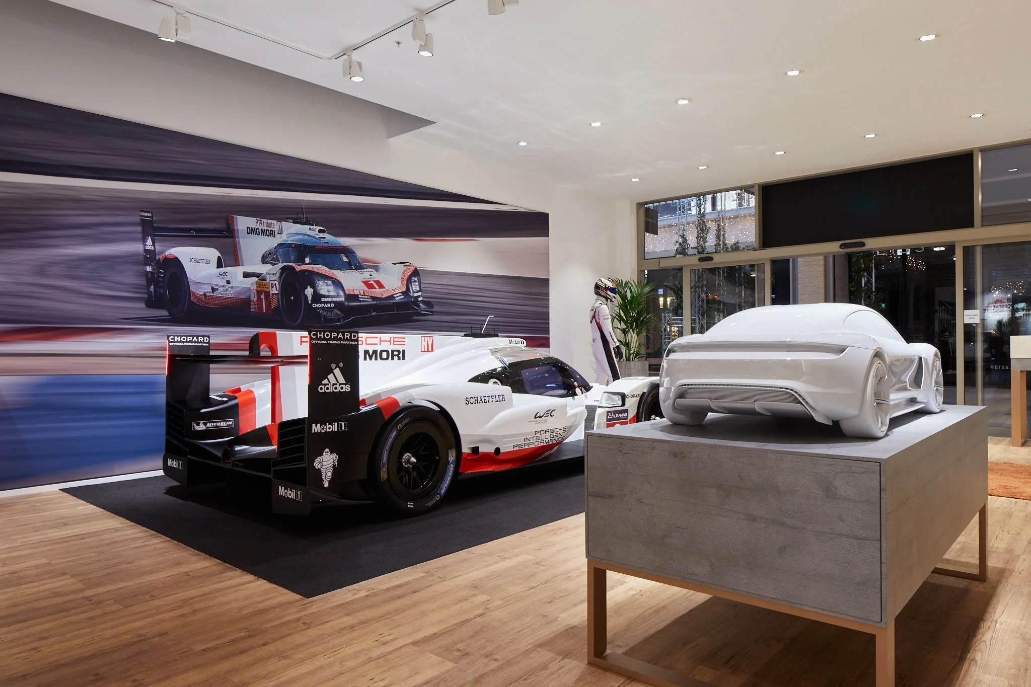 A showroom with racing cars and a sleek white concept car on display, featuring a large racing mural in the background and modern lighting overhead.