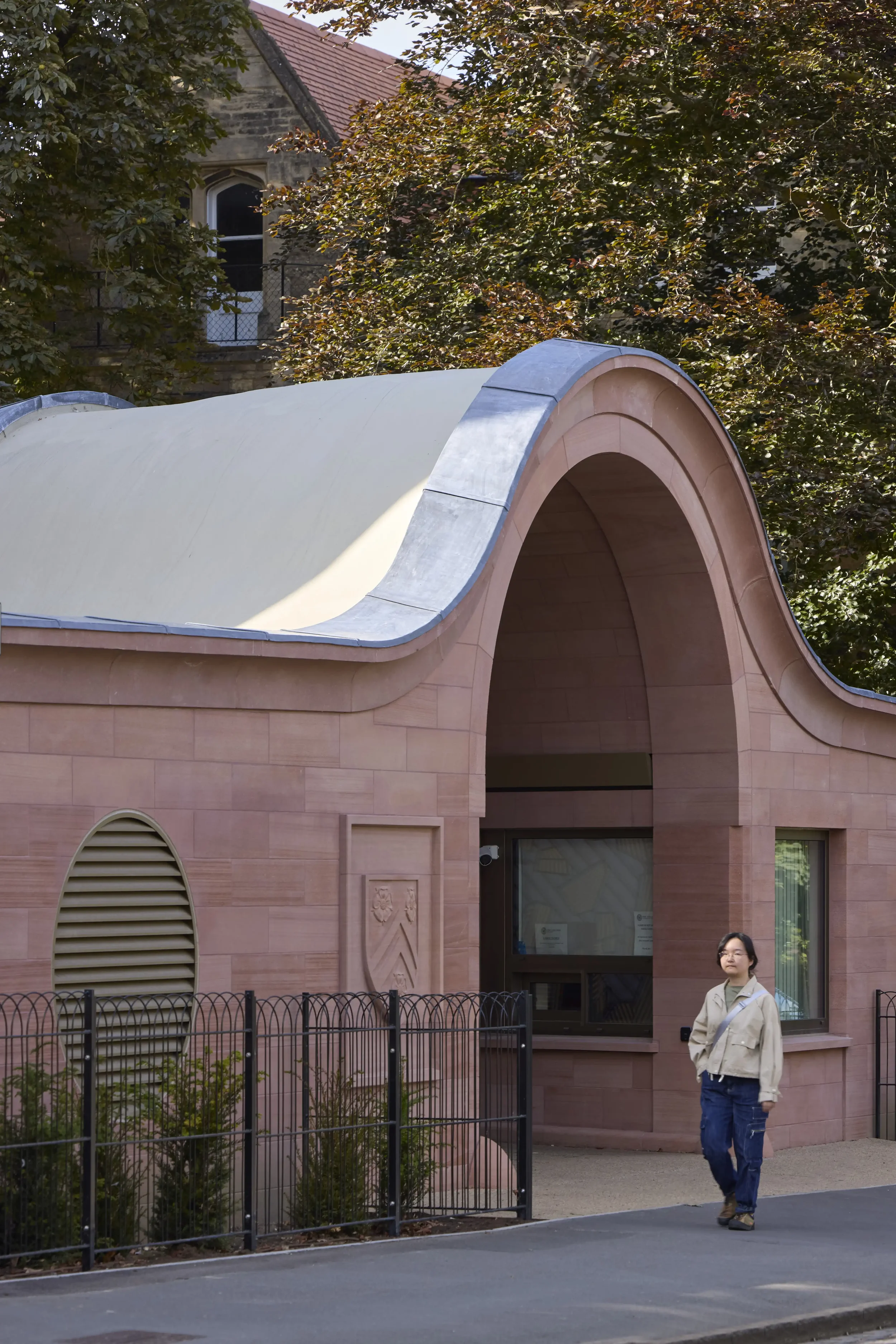 A person with glasses and light-colored jacket walking on a sidewalk in front of a pink building with a curved roof and decorative crest, surrounded by a black fence and greenery, with trees and a brick building in the background.
