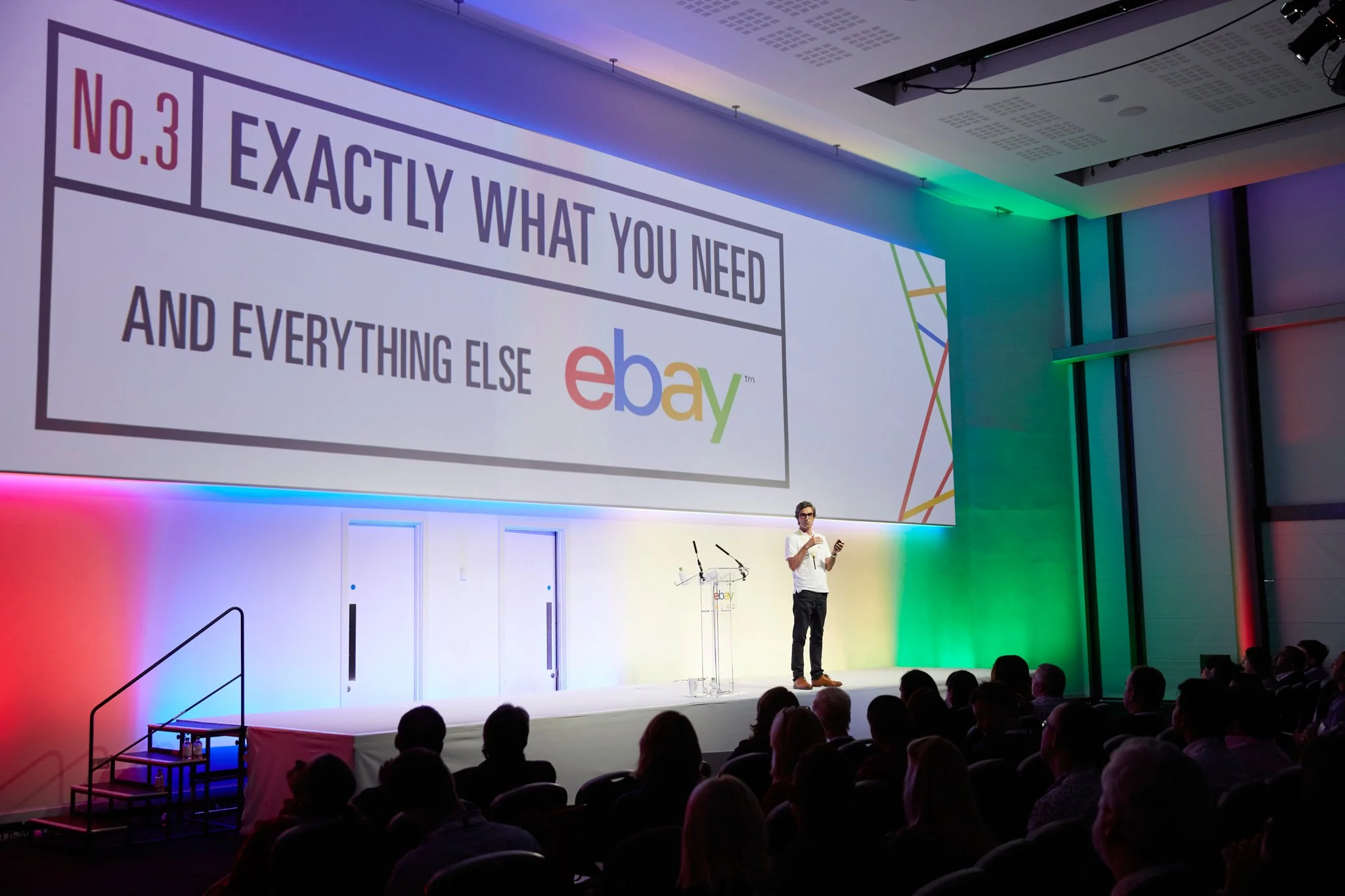 A person standing on stage giving a presentation to an audience in a conference room with colorful lighting. The large screen behind shows a slide reading 'No.3 Exactly what you need and everything else eBay'.