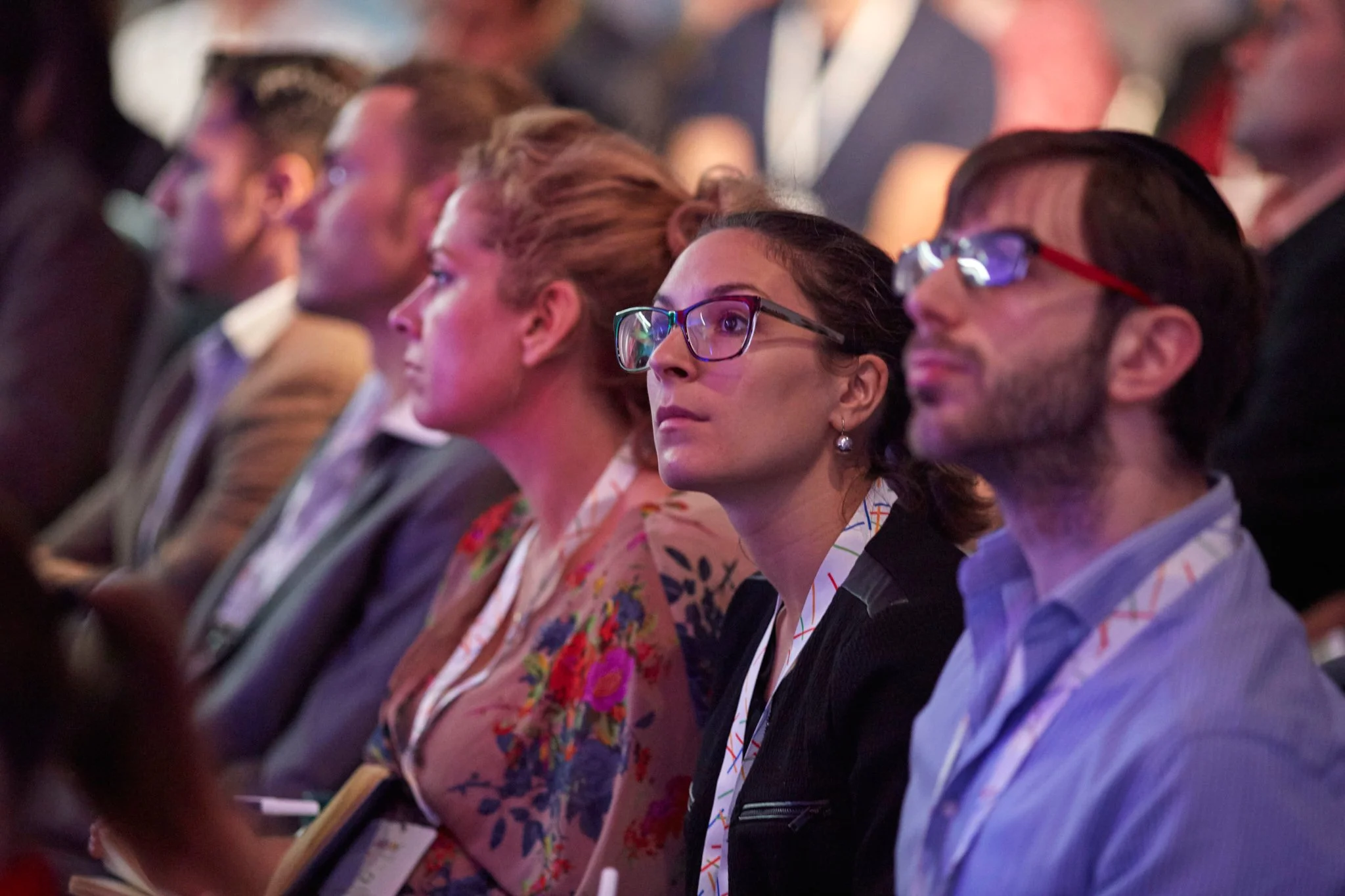 A group of people attending a conference, sitting attentively and listening, with some taking notes. The focus is on a woman with glasses and a woman with a floral blazer seated next to a man with glasses.