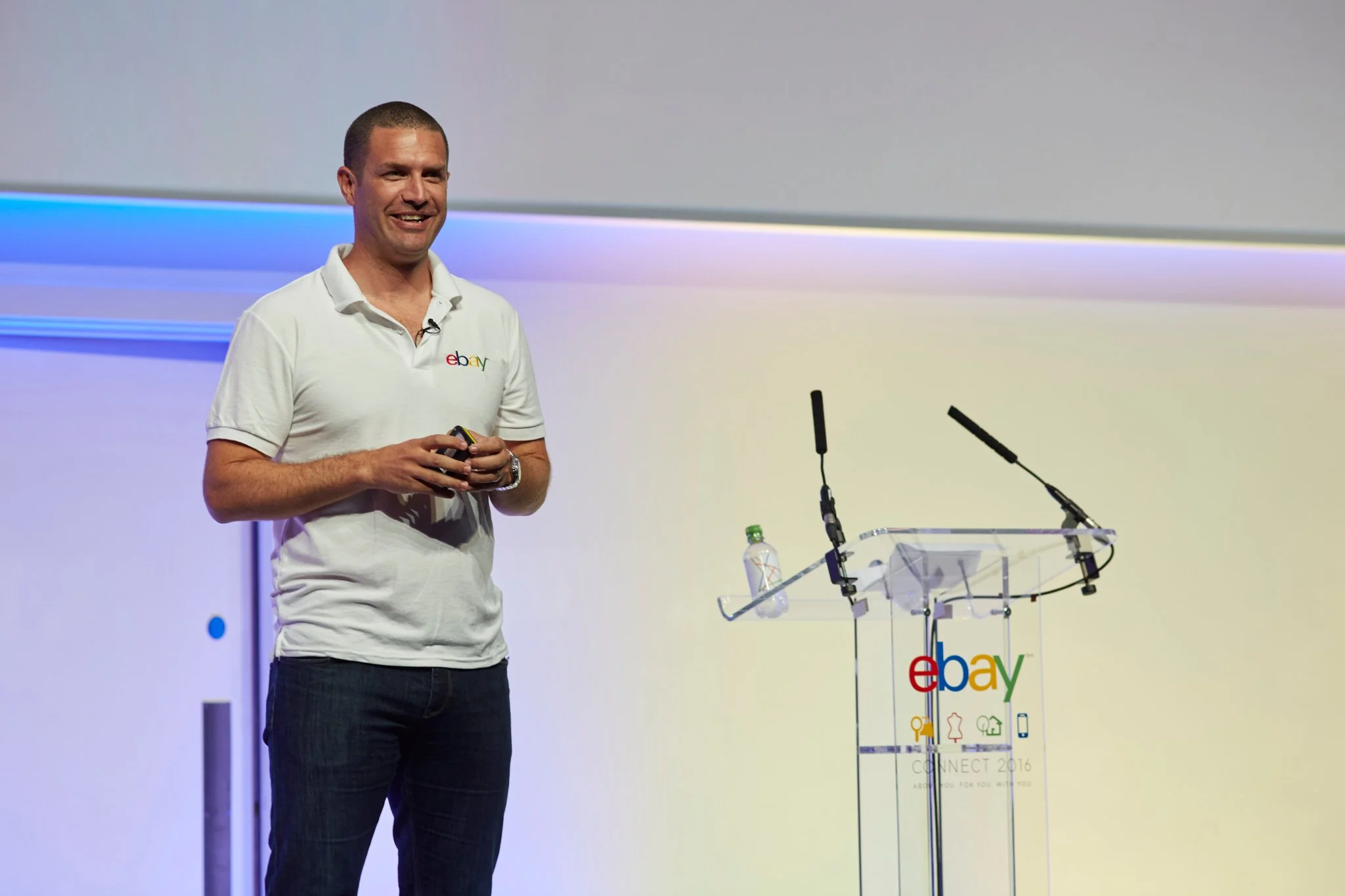 A man in a white eBay polo shirt stands on stage at eBay Connect 2016, holding a microphone, with a clear podium featuring the eBay logo and a water bottle nearby.