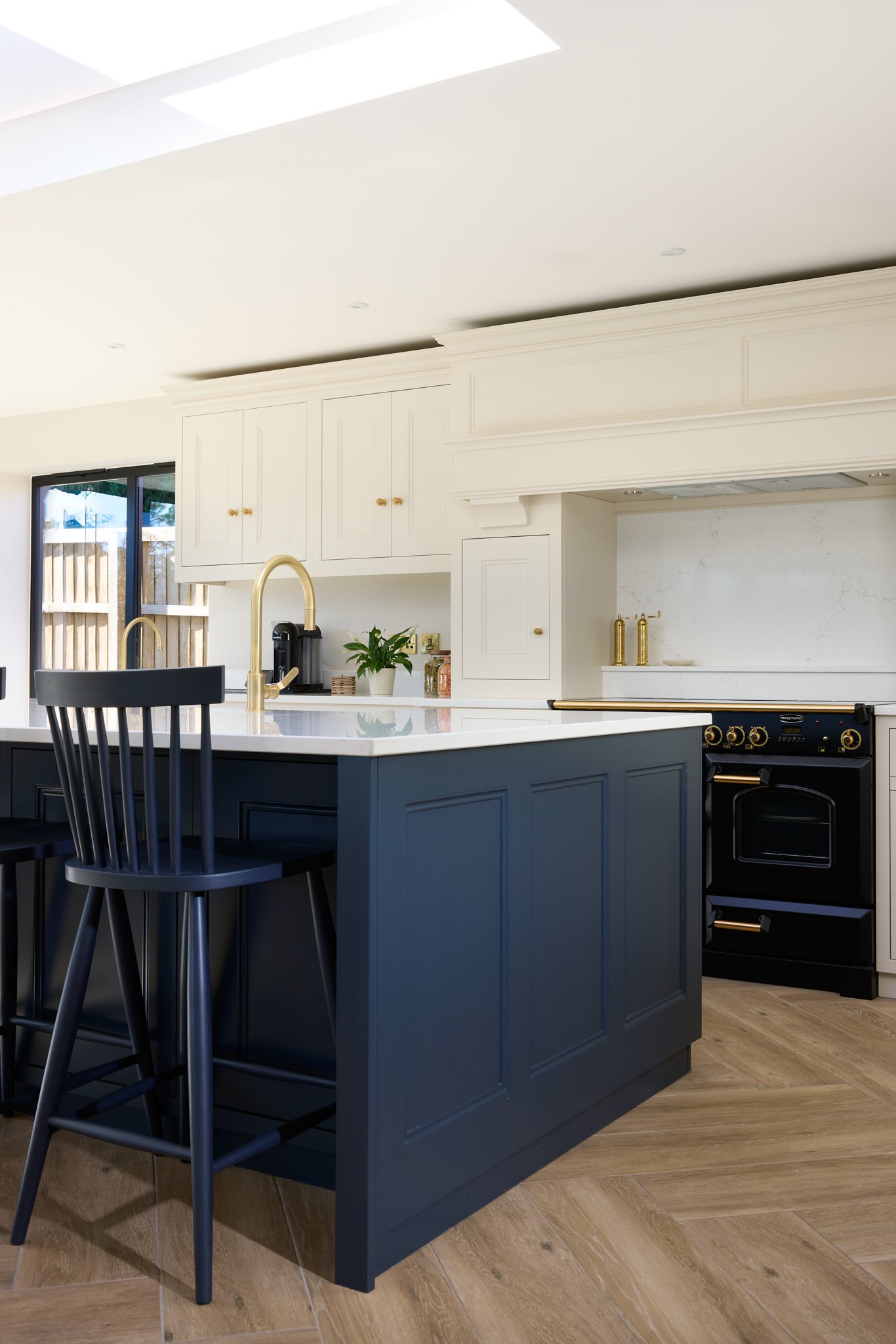 A modern kitchen with a dark blue island, black chairs, white cabinets, a gold faucet, and hardwood floors.
