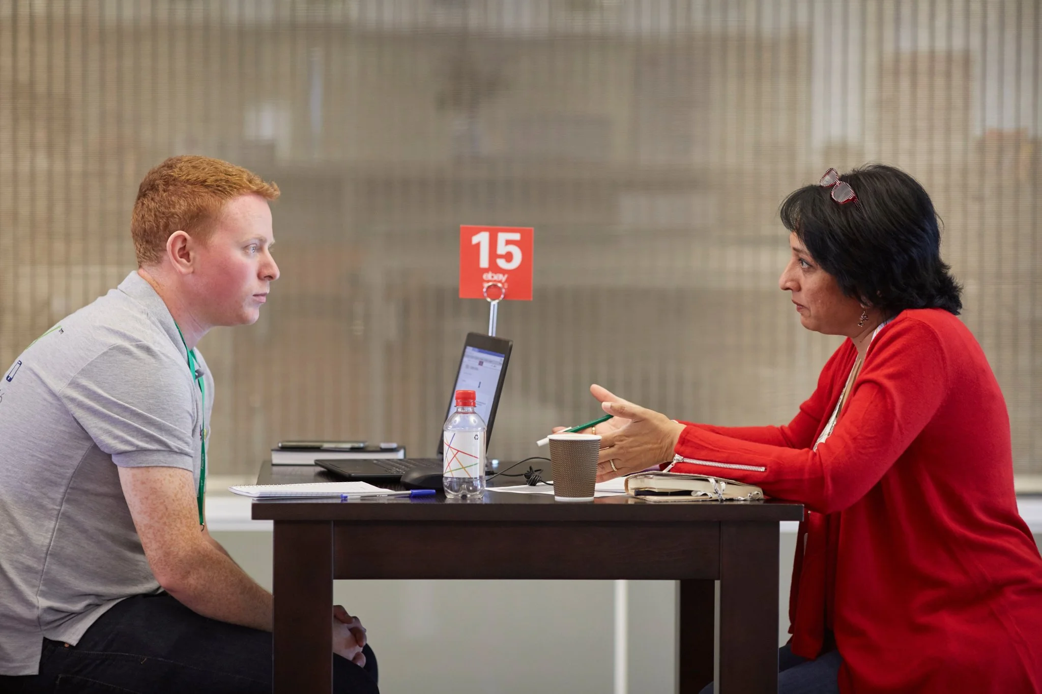 A job interview scene with a young man with red hair sitting across from a woman with black hair and glasses on her head. They are seated at a table with a laptop, notepad, and water bottle, engaged in conversation. A red sign with the number 15 is o